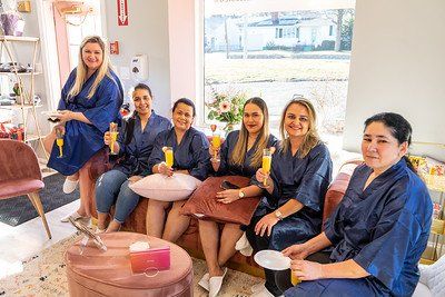 Group of women in blue robes holding drinks, smiling, gathered indoors by a window.
