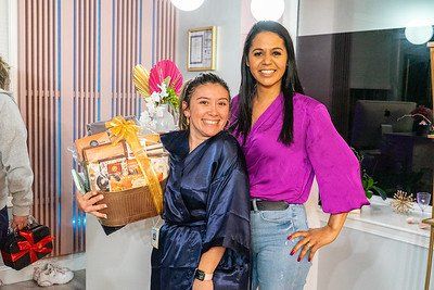 Two women smiling, one in a robe holding a gift basket, the other in purple top.