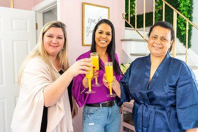 Three women in a room, toasting with orange drinks. Pink walls, gold accents, staircase with greenery.