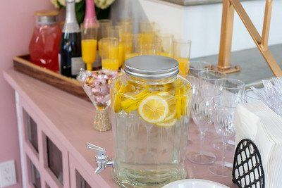 Pink table with drinks: lemon water dispenser, mimosas, juice, champagne, and glassware.