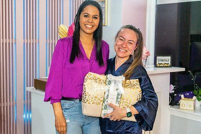 Two women smiling, holding a gift bag, in a colorful room. One wears a purple shirt, the other a robe.