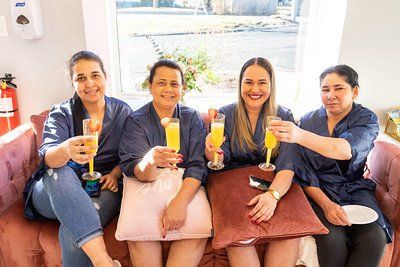 Four women in blue robes on a pink couch, toasting with mimosas near a window.