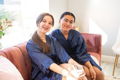 Two young women in blue robes smiling on a pink couch.