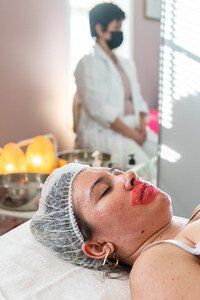 Woman with red lip mask, eyes closed, at spa; staff in background wearing a mask.