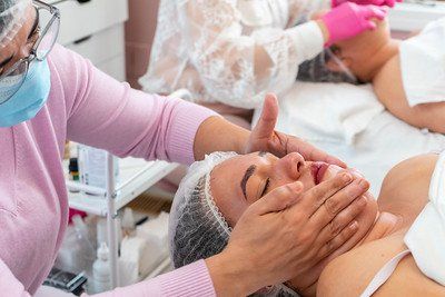 Woman receiving facial treatment at spa, hands on face, other people in background.