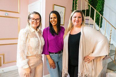 Three smiling women stand indoors; two women stand on either side of a woman wearing a purple top.