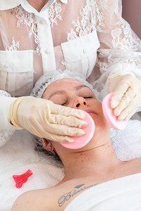 Woman receiving facial treatment with pink pads; esthetician in white lace shirt and gloves.