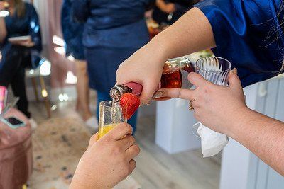 Hands pouring a bubbly pink drink into a glass with a strawberry, in a room with people.
