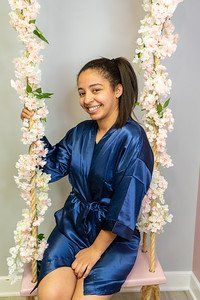 Woman in navy satin robe smiles, sitting on a pink swing adorned with flowers.