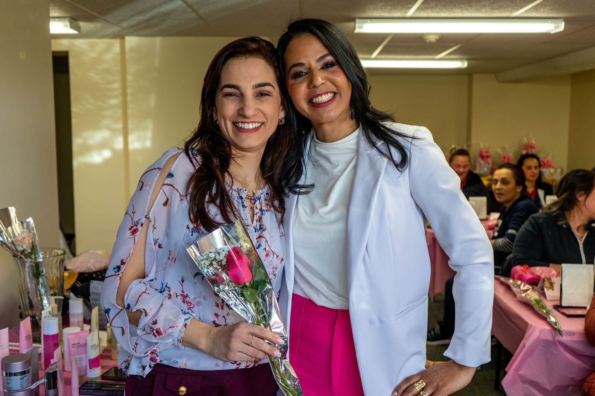 Two smiling women, one holding a rose, pose together in a room decorated in pink.