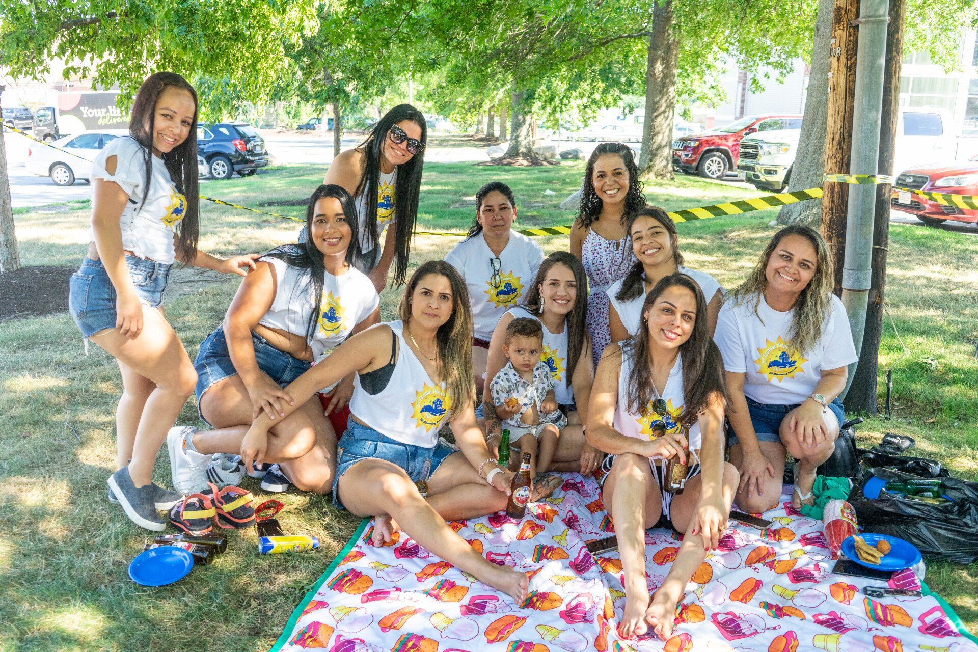 Group of women and a child picnicking in a park. They wear white t-shirts and denim shorts.
