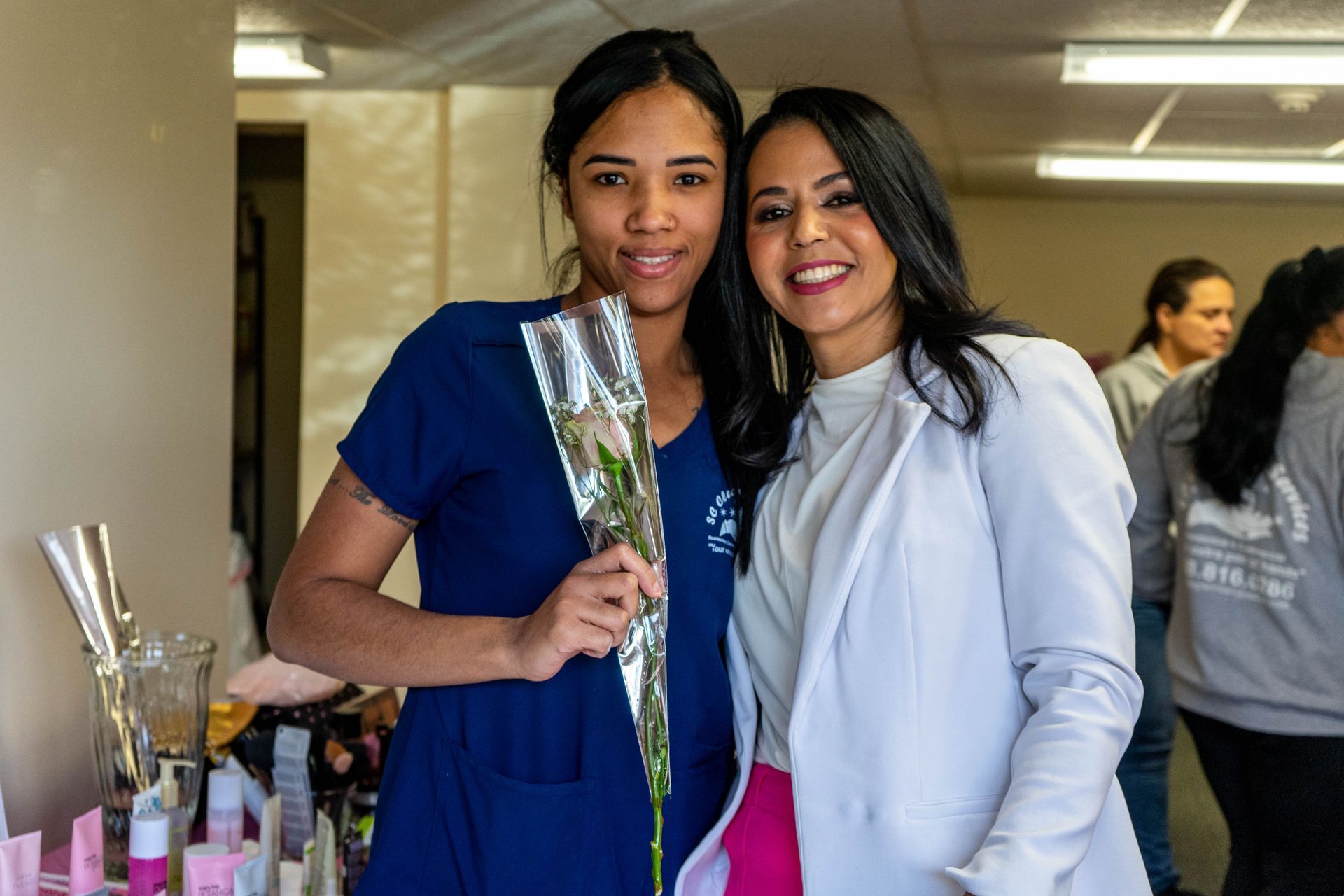 Two women smiling, one holding a rose. Woman on left in blue scrubs, the other in a white coat. Inside setting.