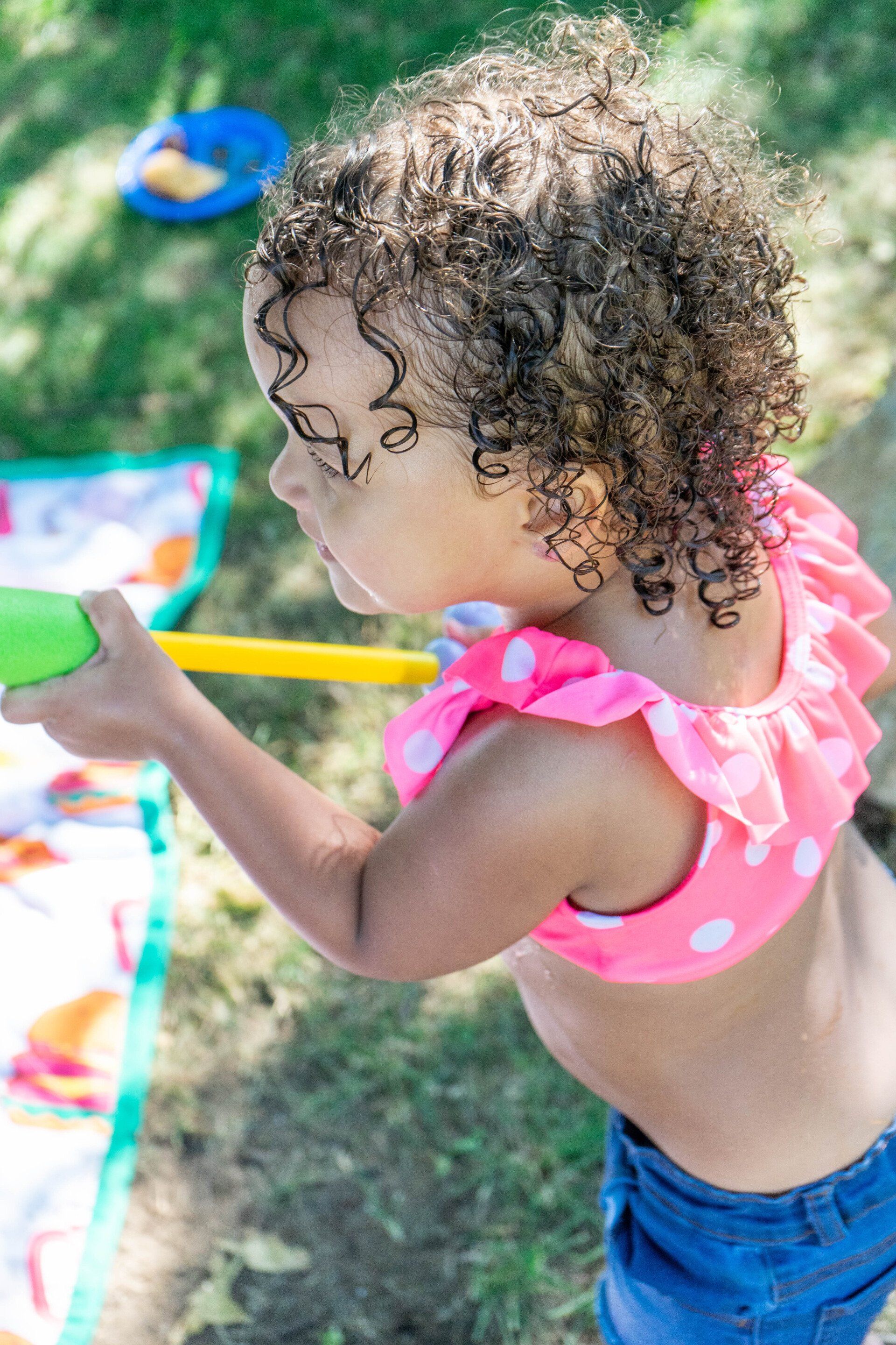 Young child with curly hair aiming a water gun, wearing a pink polka dot swim top and blue shorts outside.