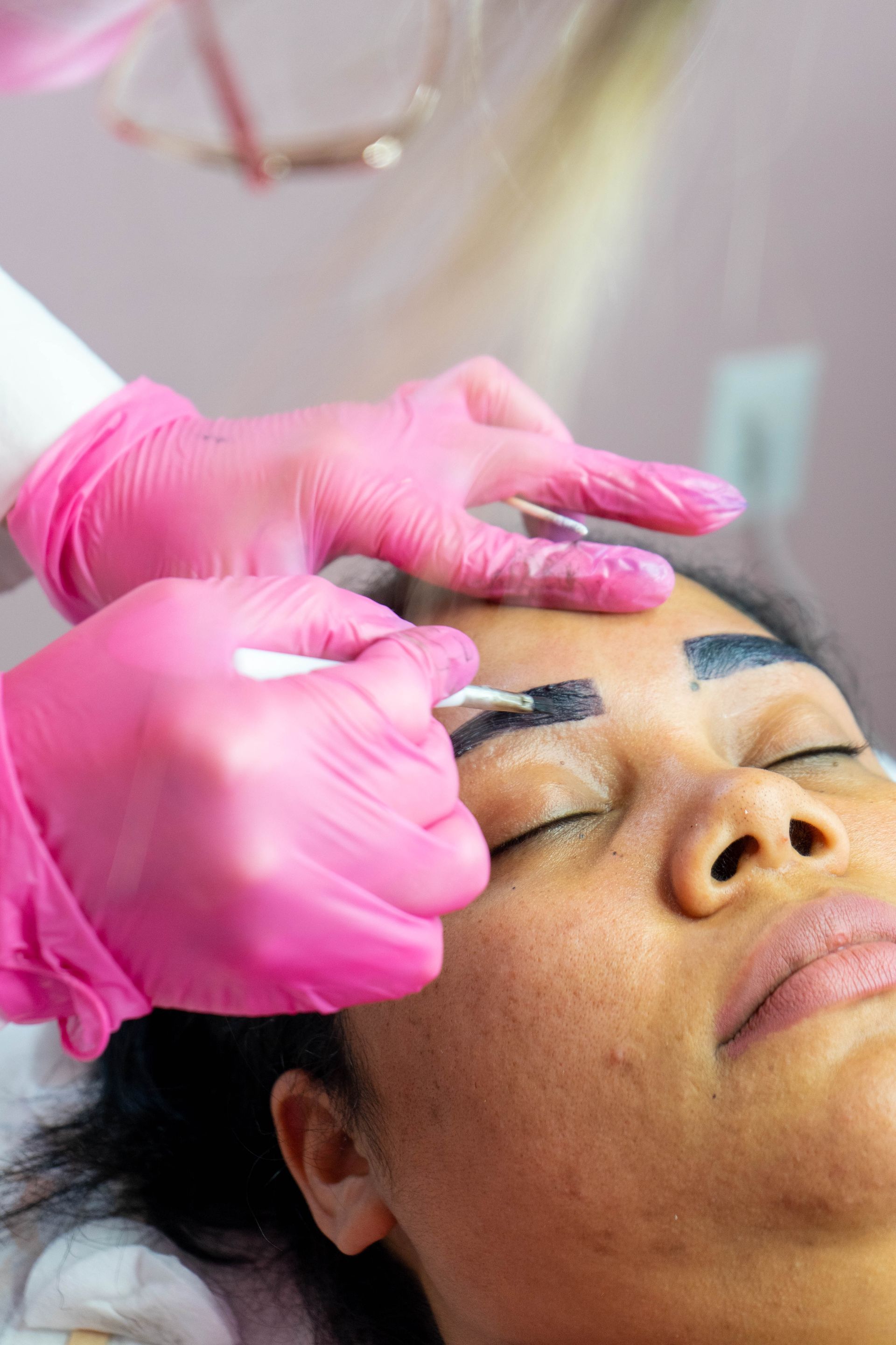 Person receiving eyebrow treatment; beautician applying pigment with pink gloves in a salon.