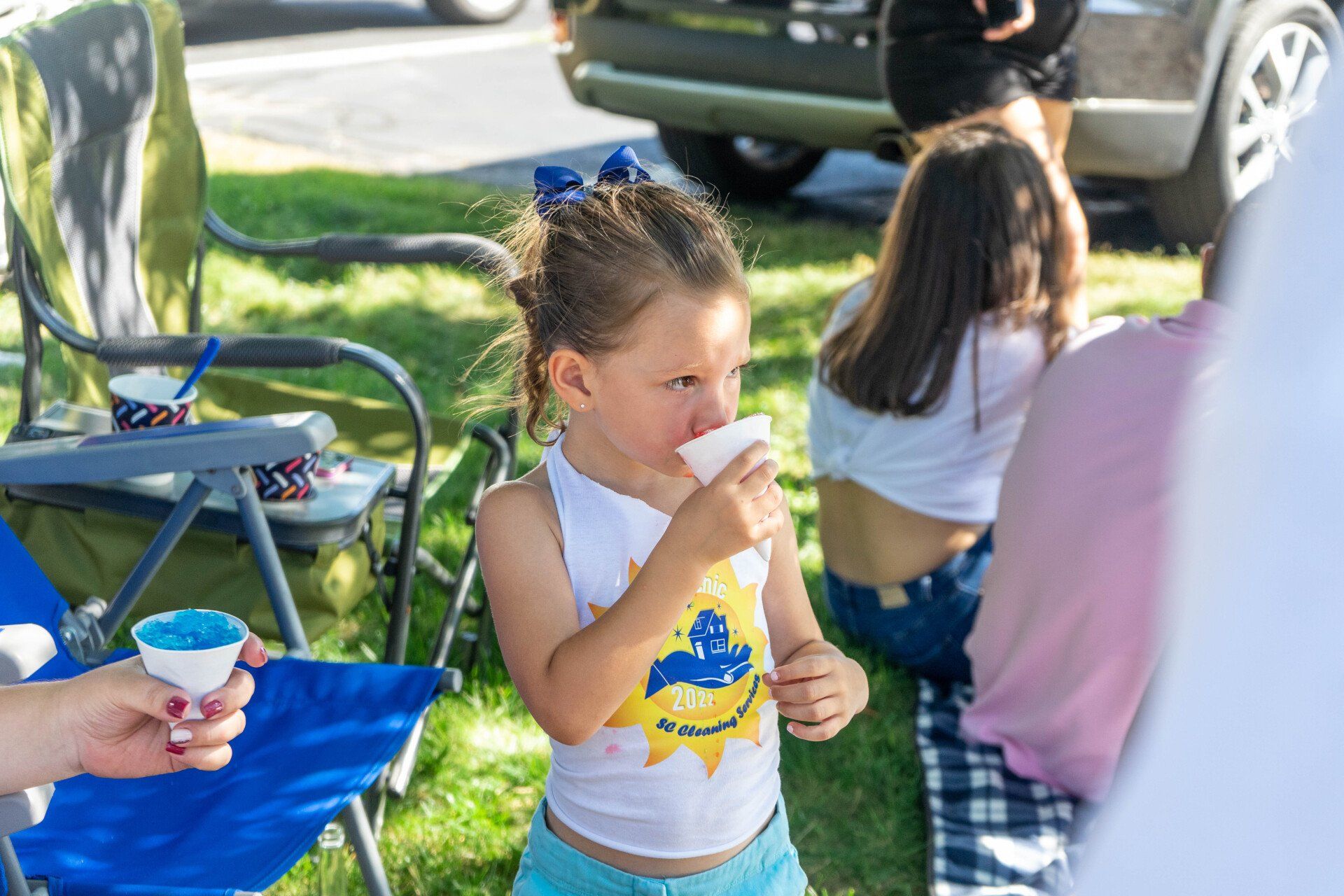 Girl drinks a blue ice treat at an outdoor event, with people and cars in the background.