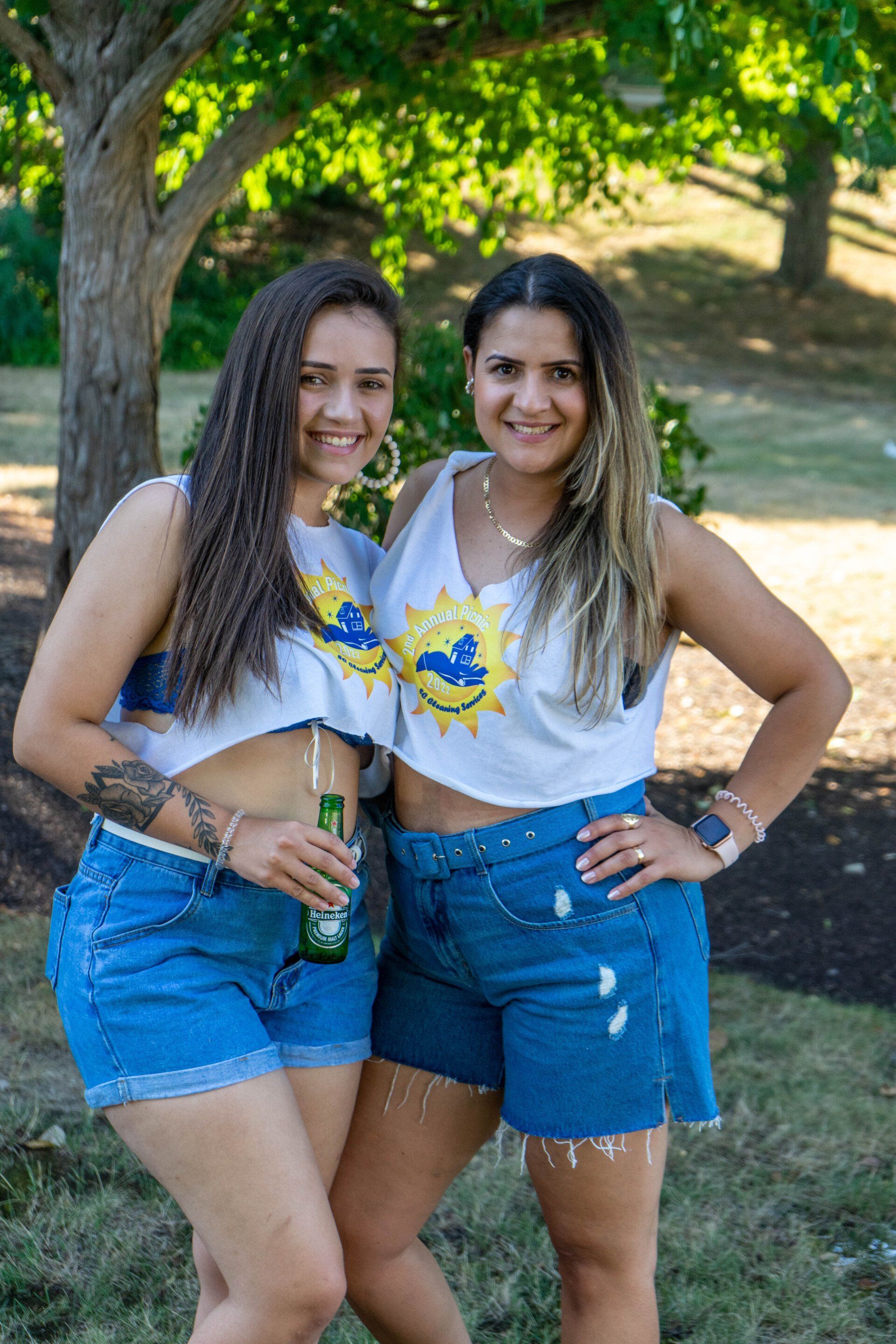 Two women in denim shorts and crop tops smile outdoors, with a tree in the background.