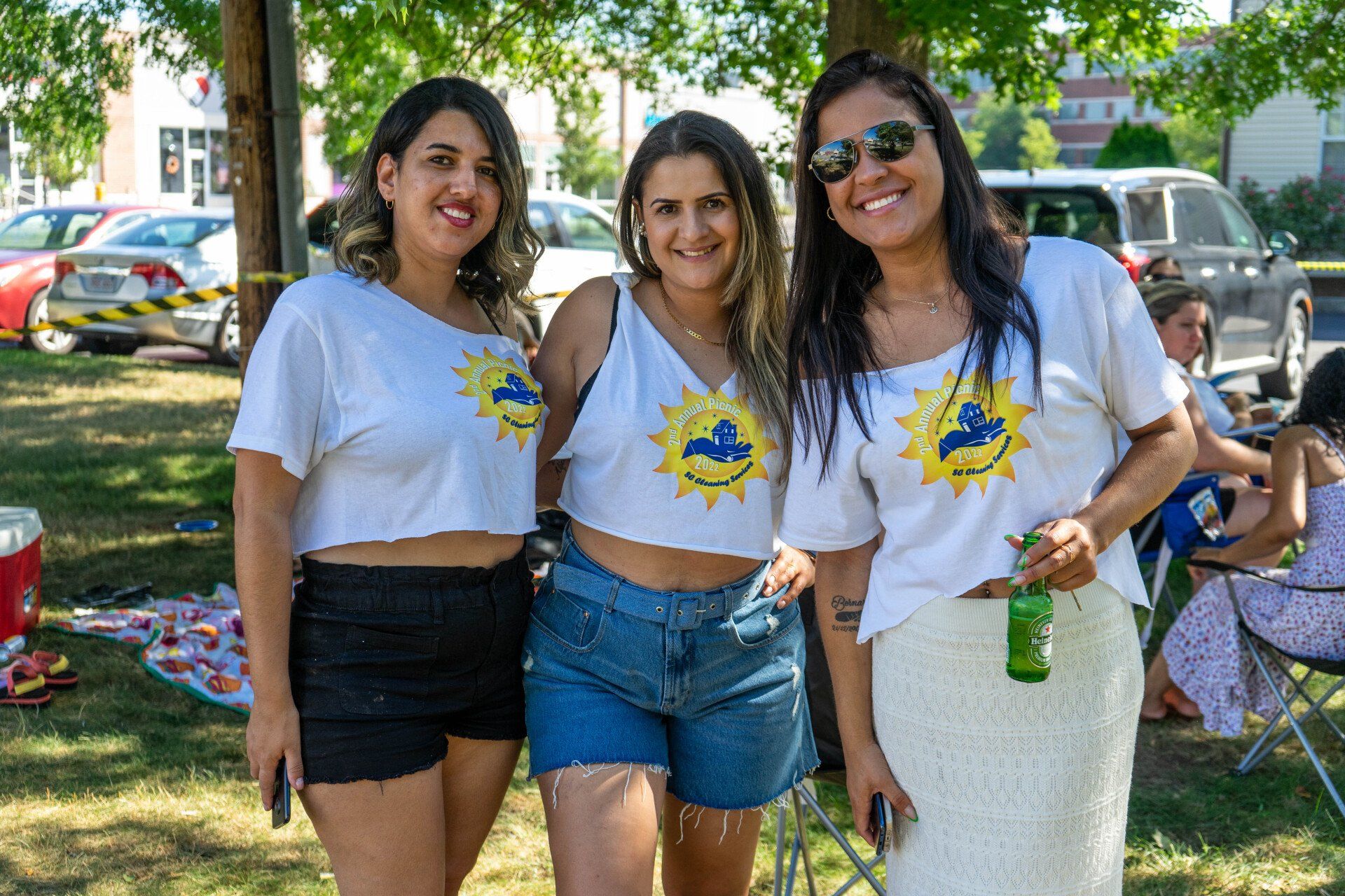 Three smiling women in white crop tops with a sun logo pose outdoors. One has sunglasses.