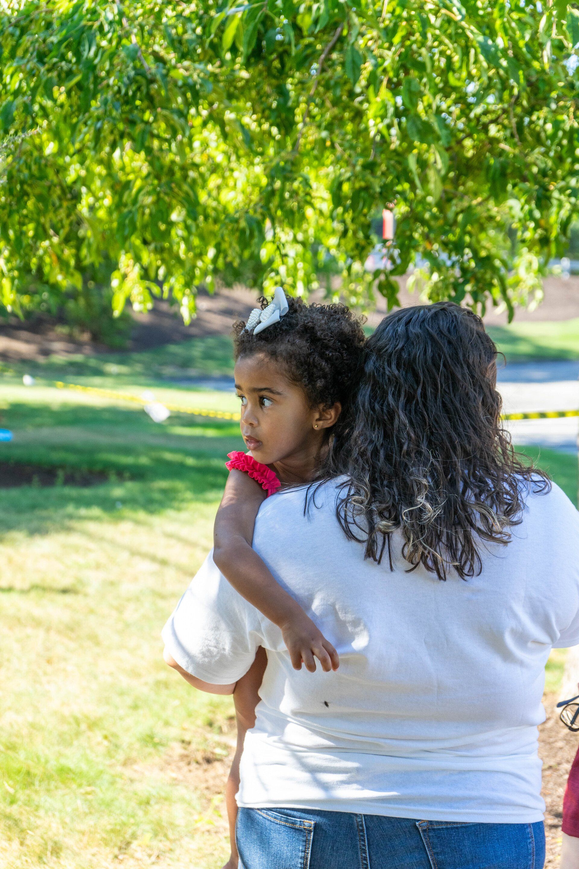 Woman in white shirt holds a young girl with curly hair outdoors, looking away.