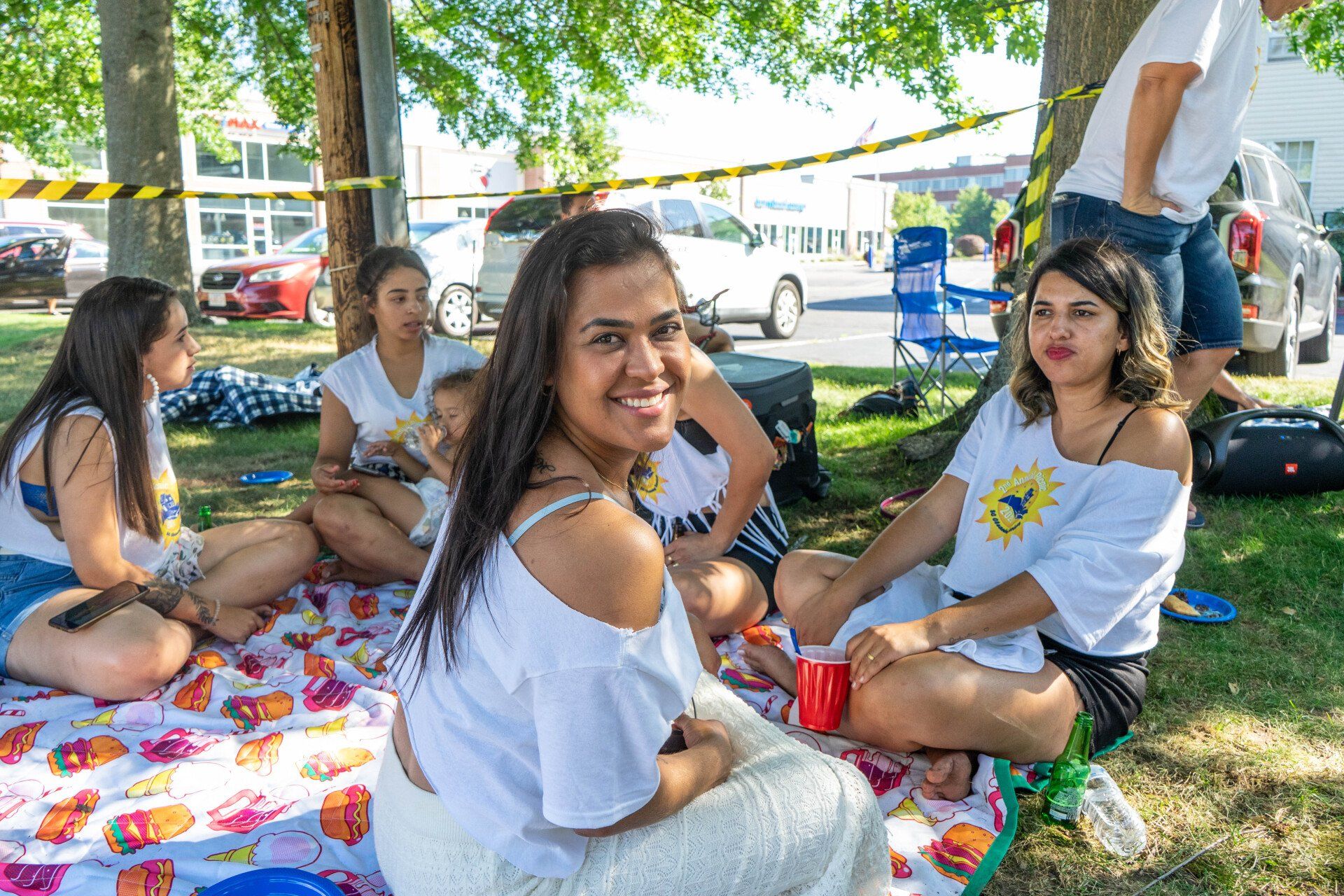 Group of people on a blanket in a park. Smiling women, one with a baby. Sunshine, cars, trees.