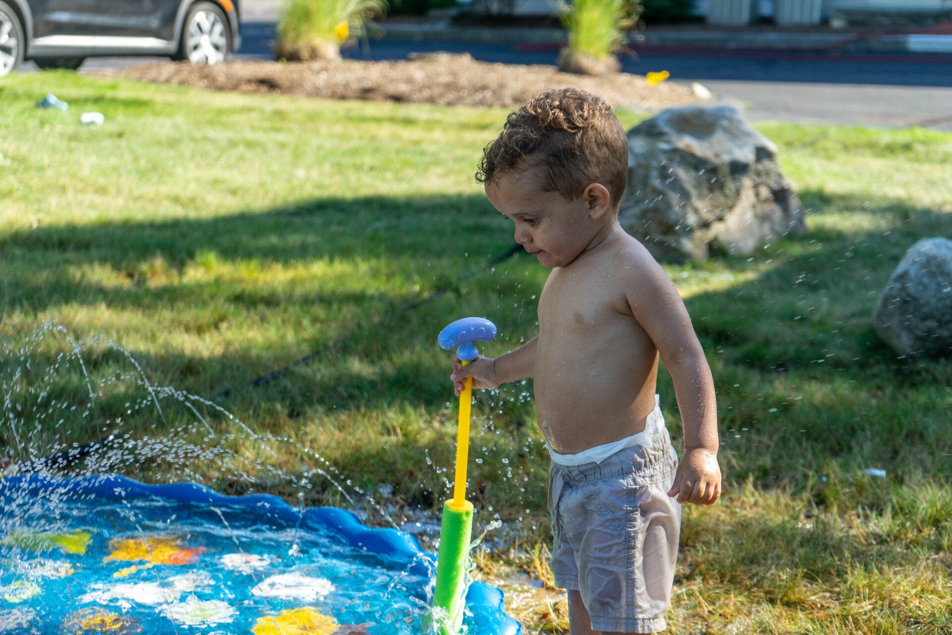 Young child plays with a water toy in a splash pad on a sunny day, surrounded by grass and rocks.
