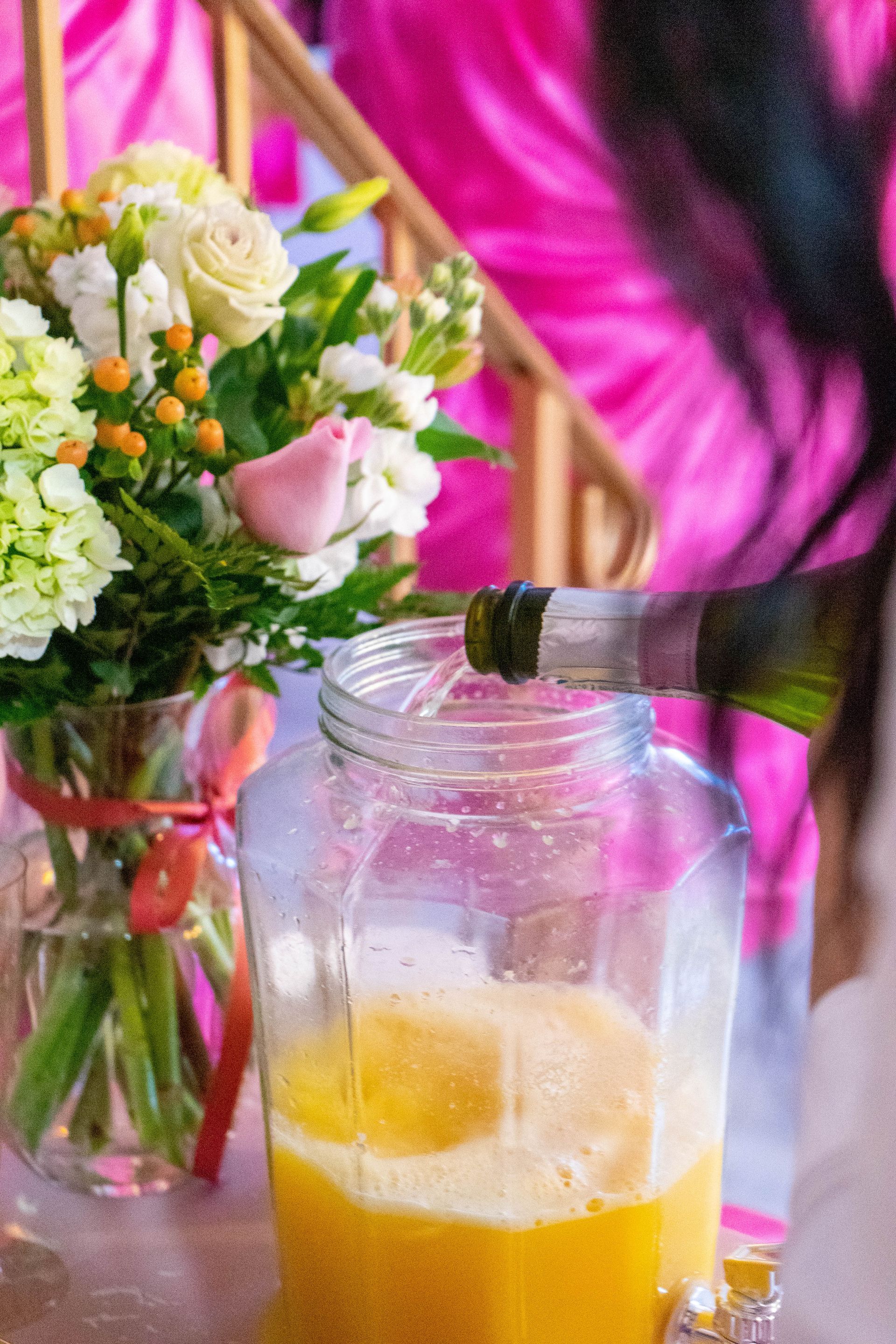 Champagne being poured into a jar of orange juice, near a flower arrangement; pink backdrop.