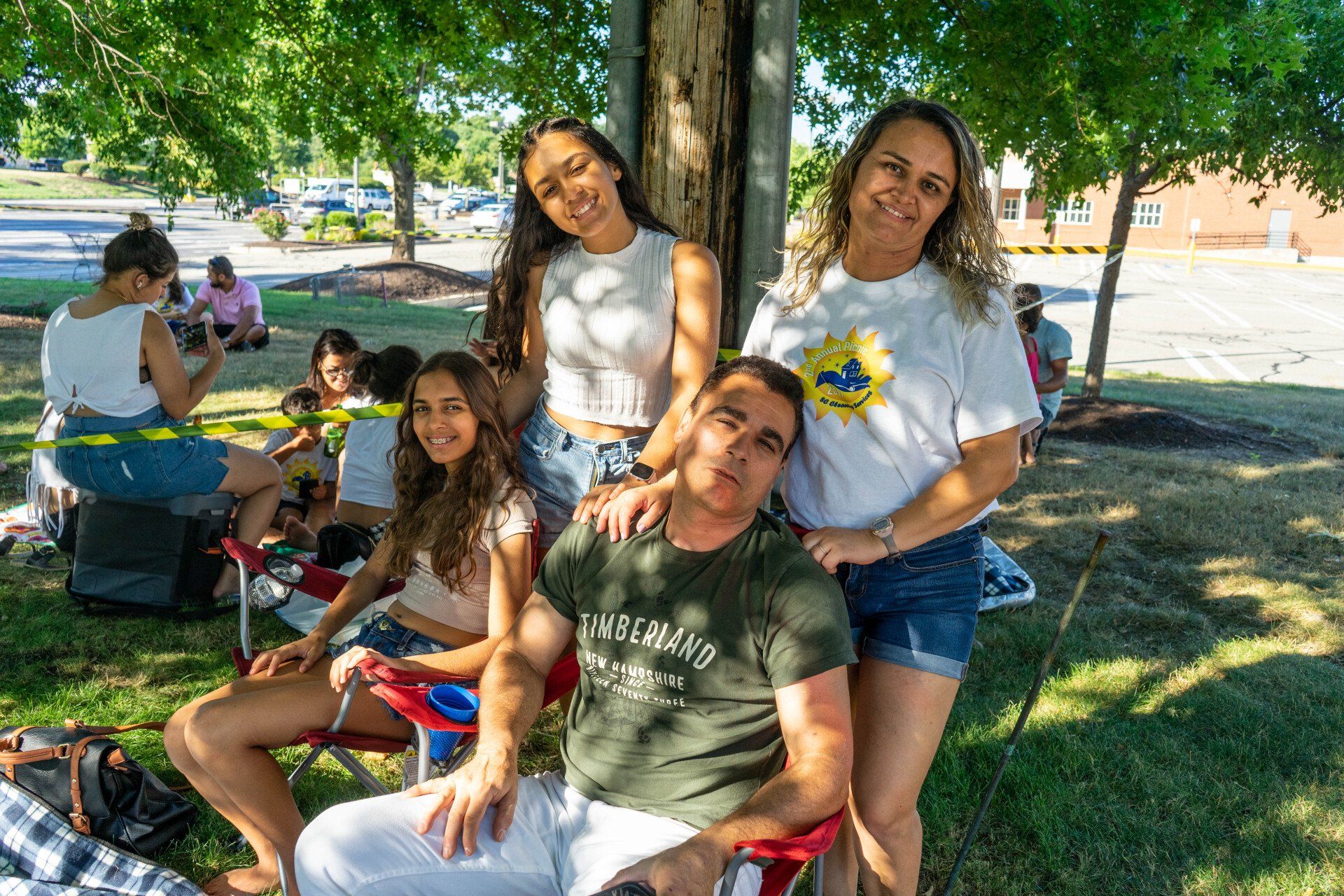 Family poses under a tree at an outdoor gathering; sunny day.