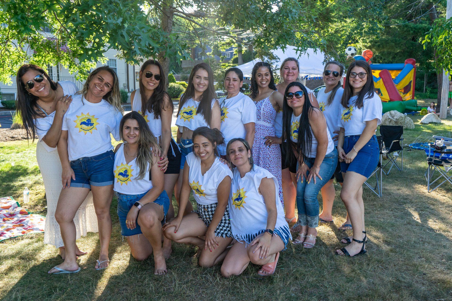 Group of women in matching white t-shirts pose outdoors. Some stand, some kneel, smiles. Green grass, sunny day.