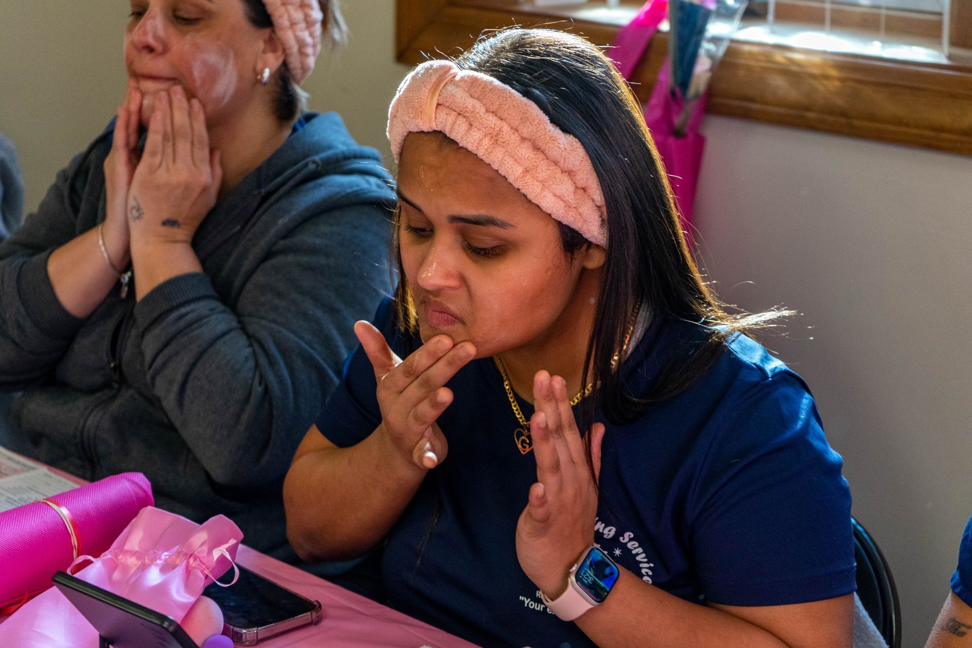 Two women applying facial cream at a table. One in a blue shirt, the other in gray, both with headbands.