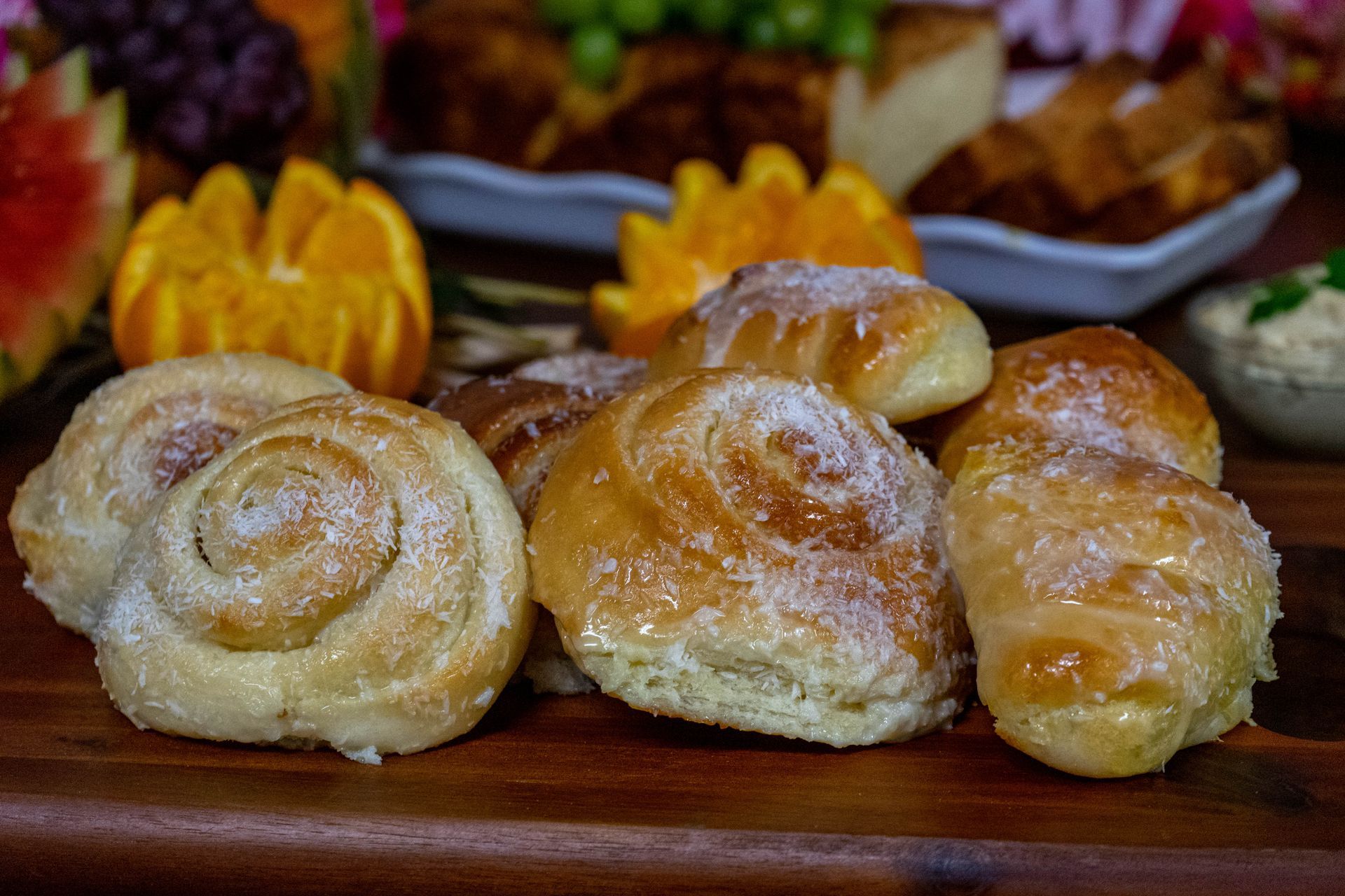 Sugar-coated cinnamon rolls on a wooden surface, with fruit and other baked goods blurred in the background.