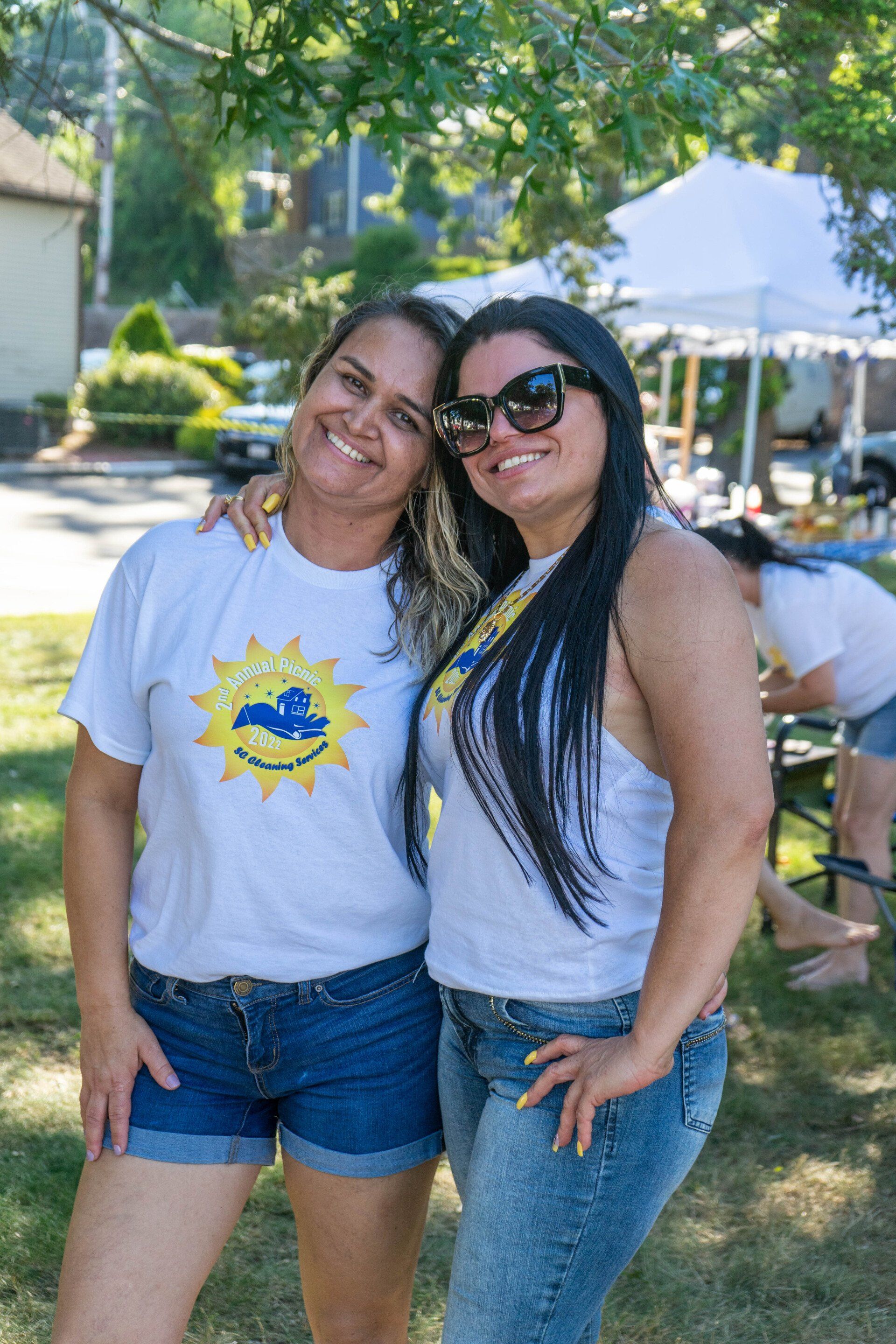 Two women smiling, posing outdoors. One in white tee, jean shorts. Other in sunglasses, white tank, jeans.