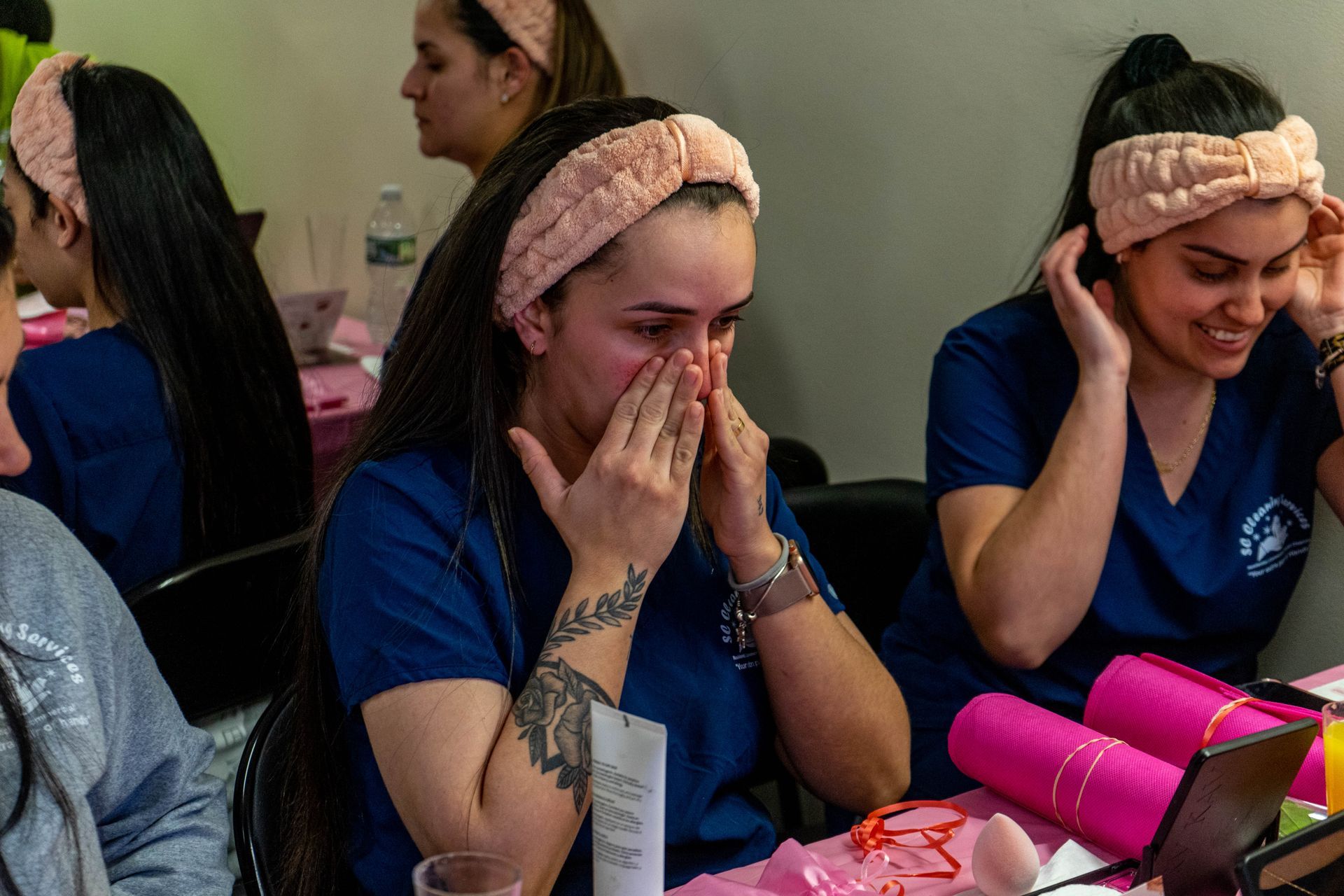 Women in blue scrubs with pink headbands, some with hands on faces, in a room with mirrors and makeup.