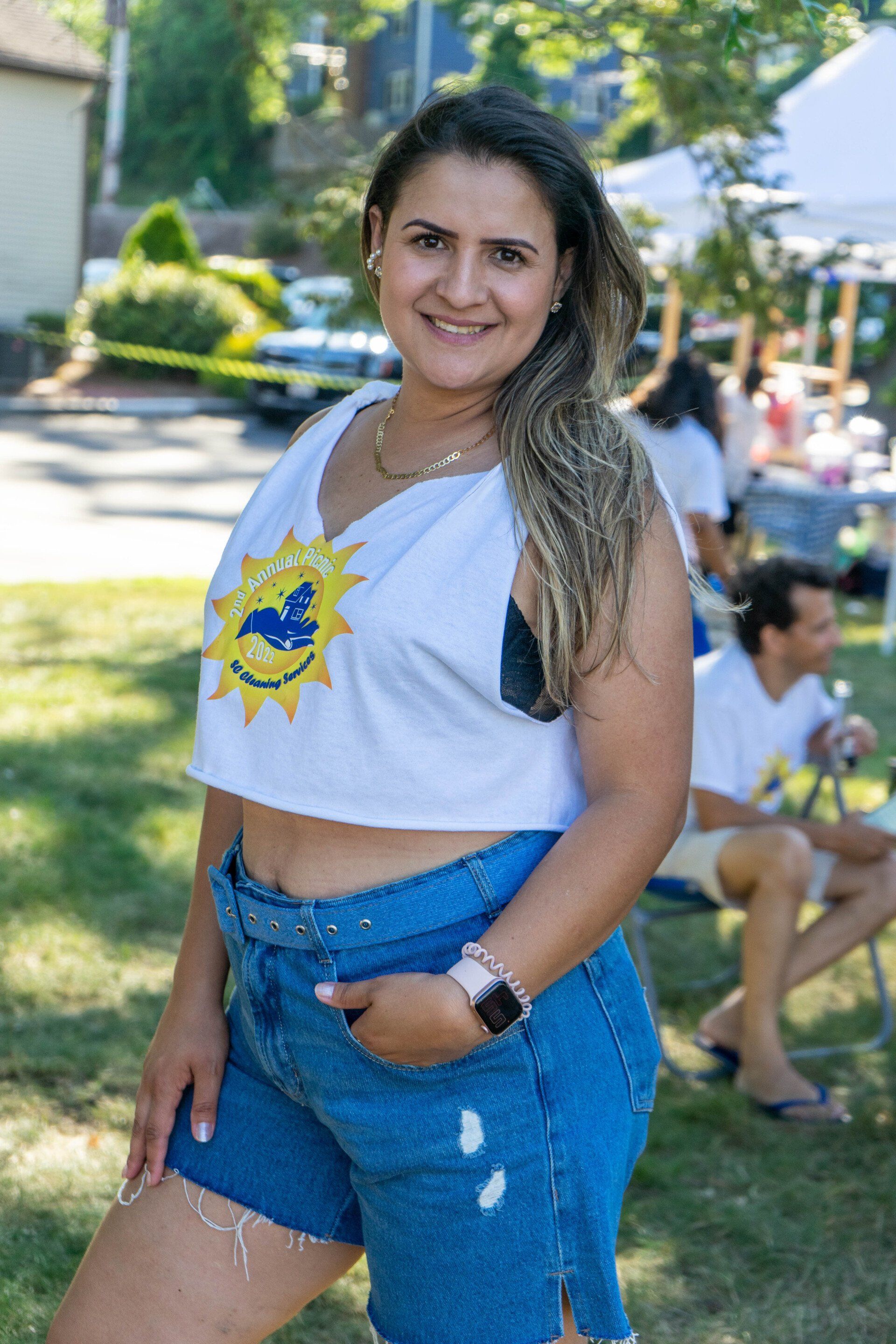 Woman smiling, outdoors, wearing a white crop top with a logo and denim shorts.