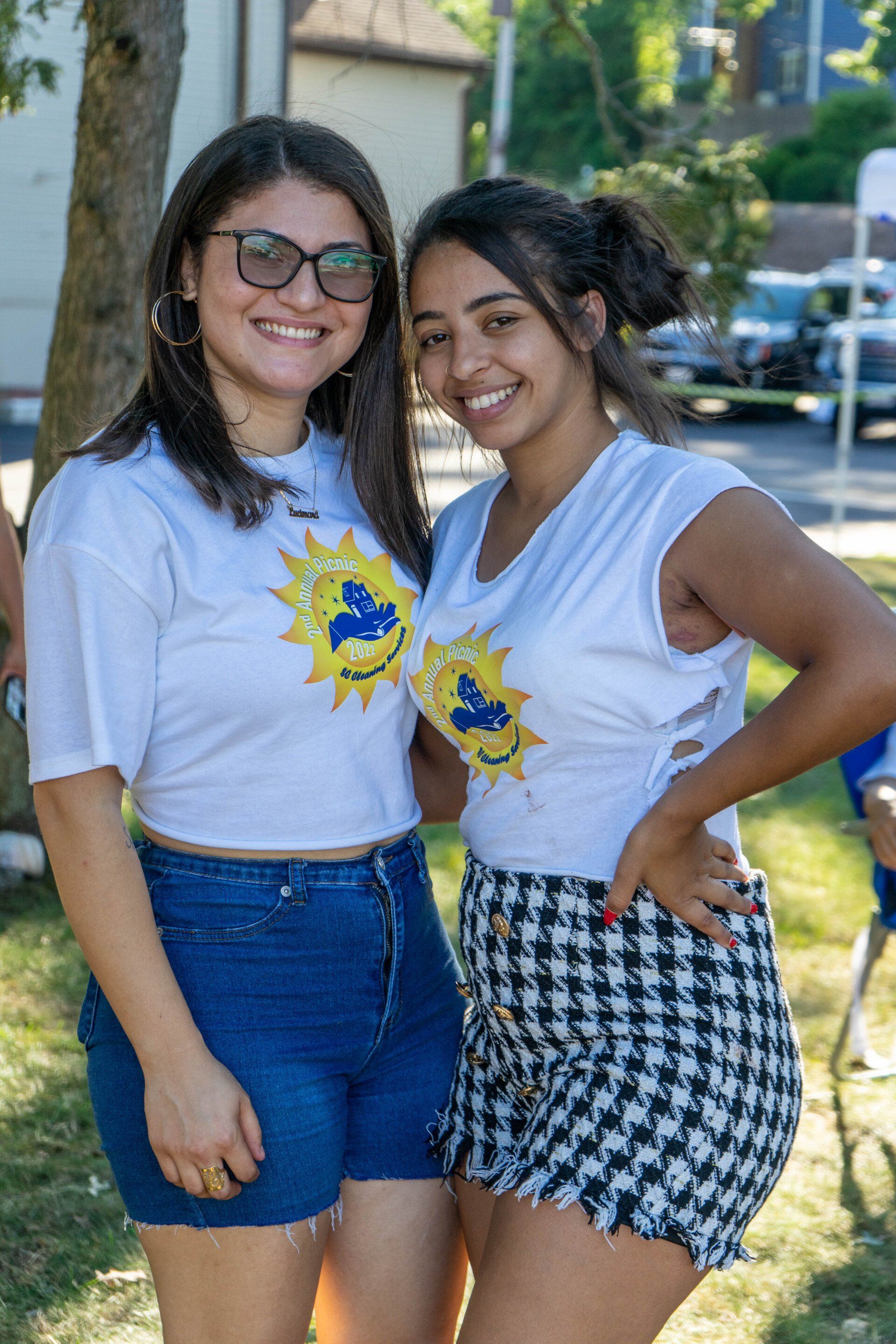 Two young women smiling outdoors, wearing white shirts with a sun design and denim shorts/skirt.