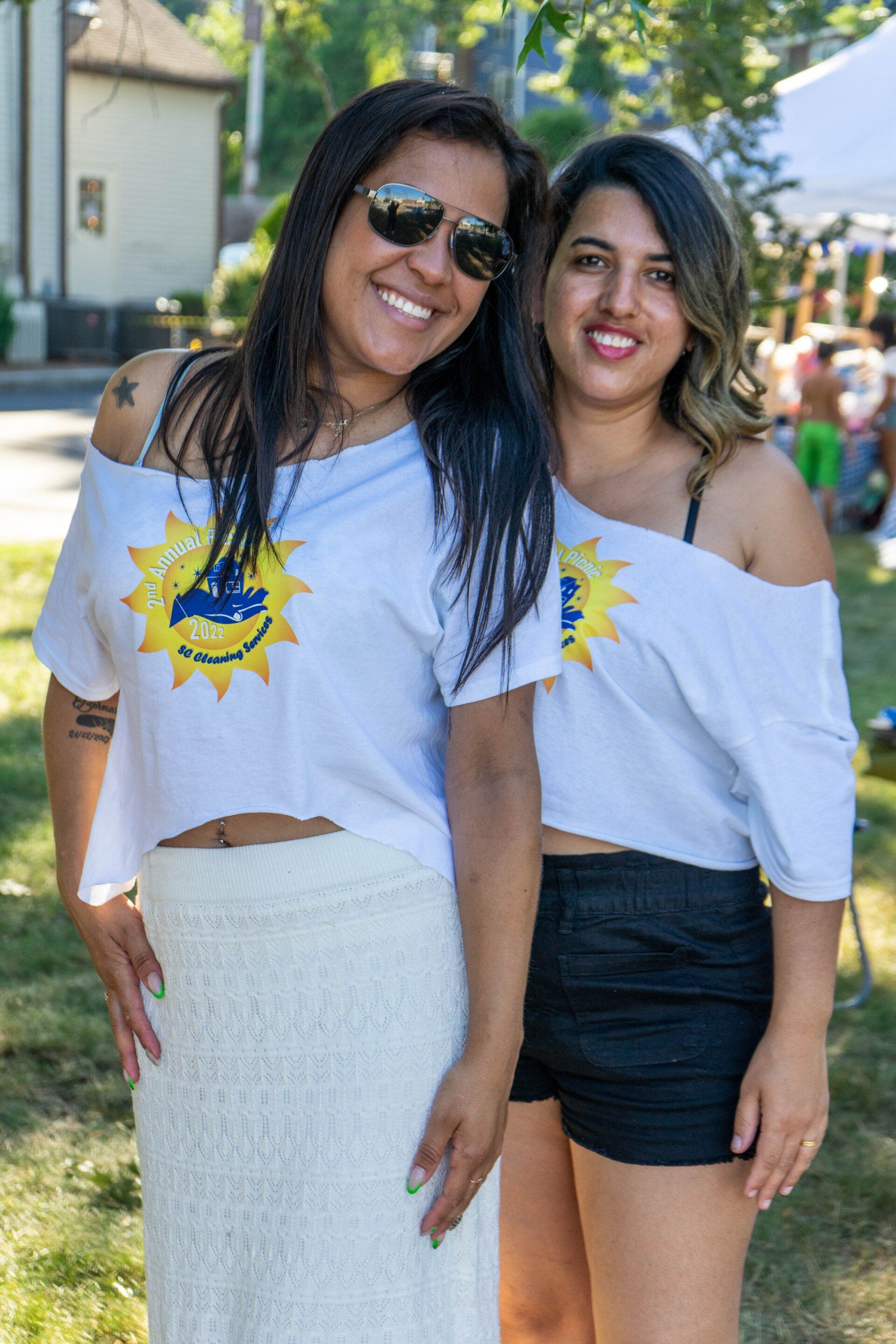 Two smiling women in white off-the-shoulder tops, outside on a sunny day; one wears a white skirt, the other black shorts.