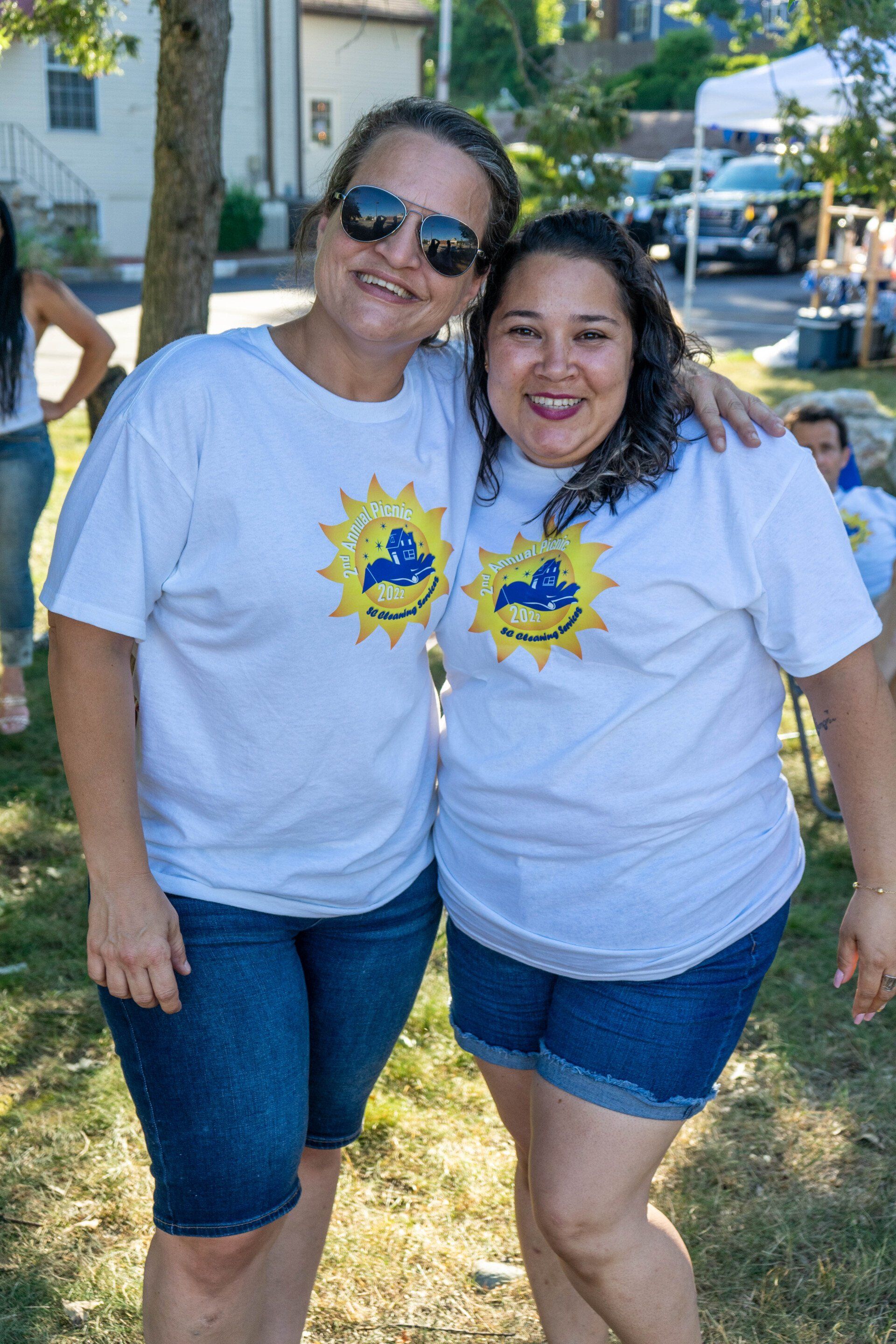 Two women smiling, wearing matching white shirts with a sun logo and jean shorts, outside.