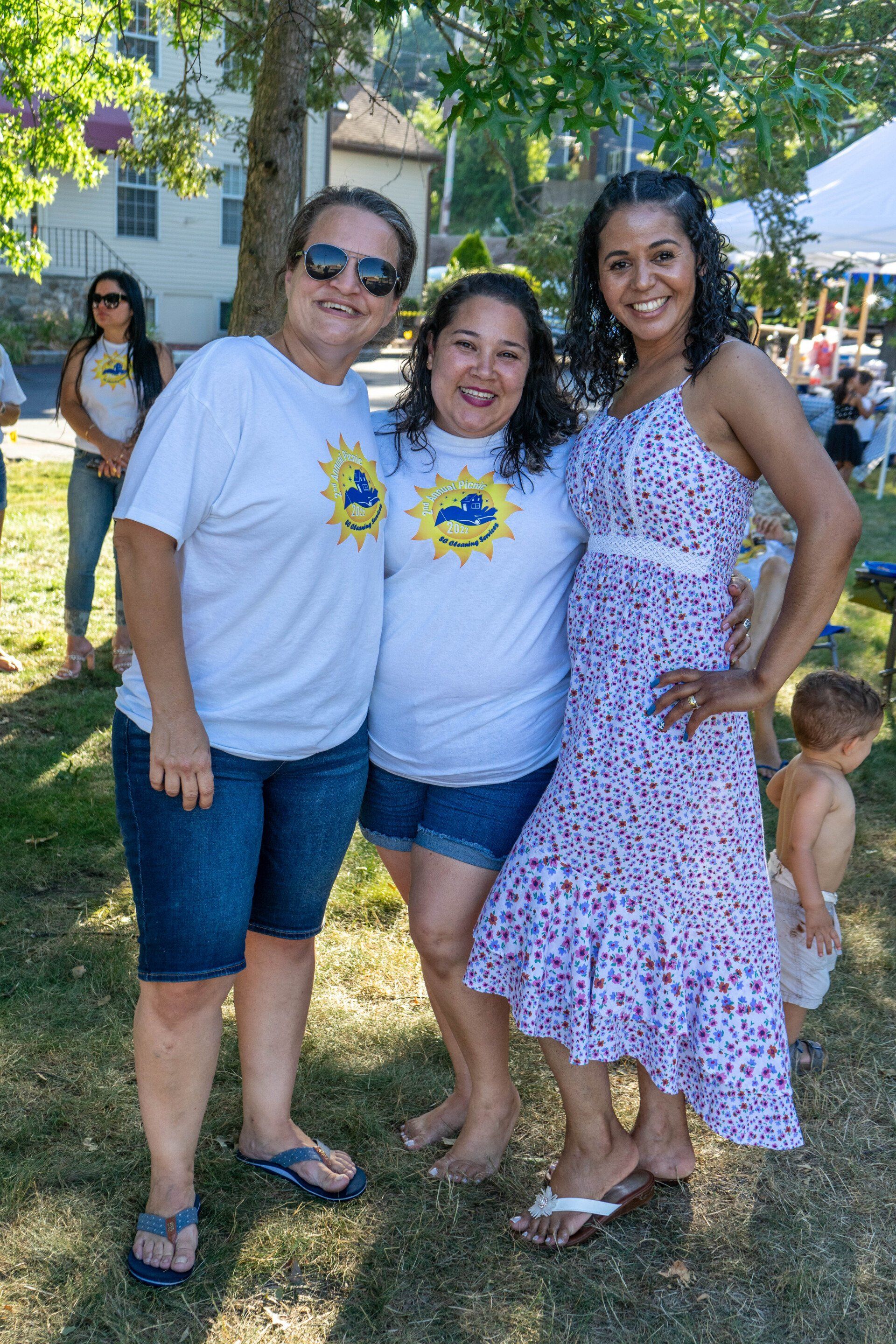 Three women smiling outdoors. One in a sundress, two in matching shirts and shorts.