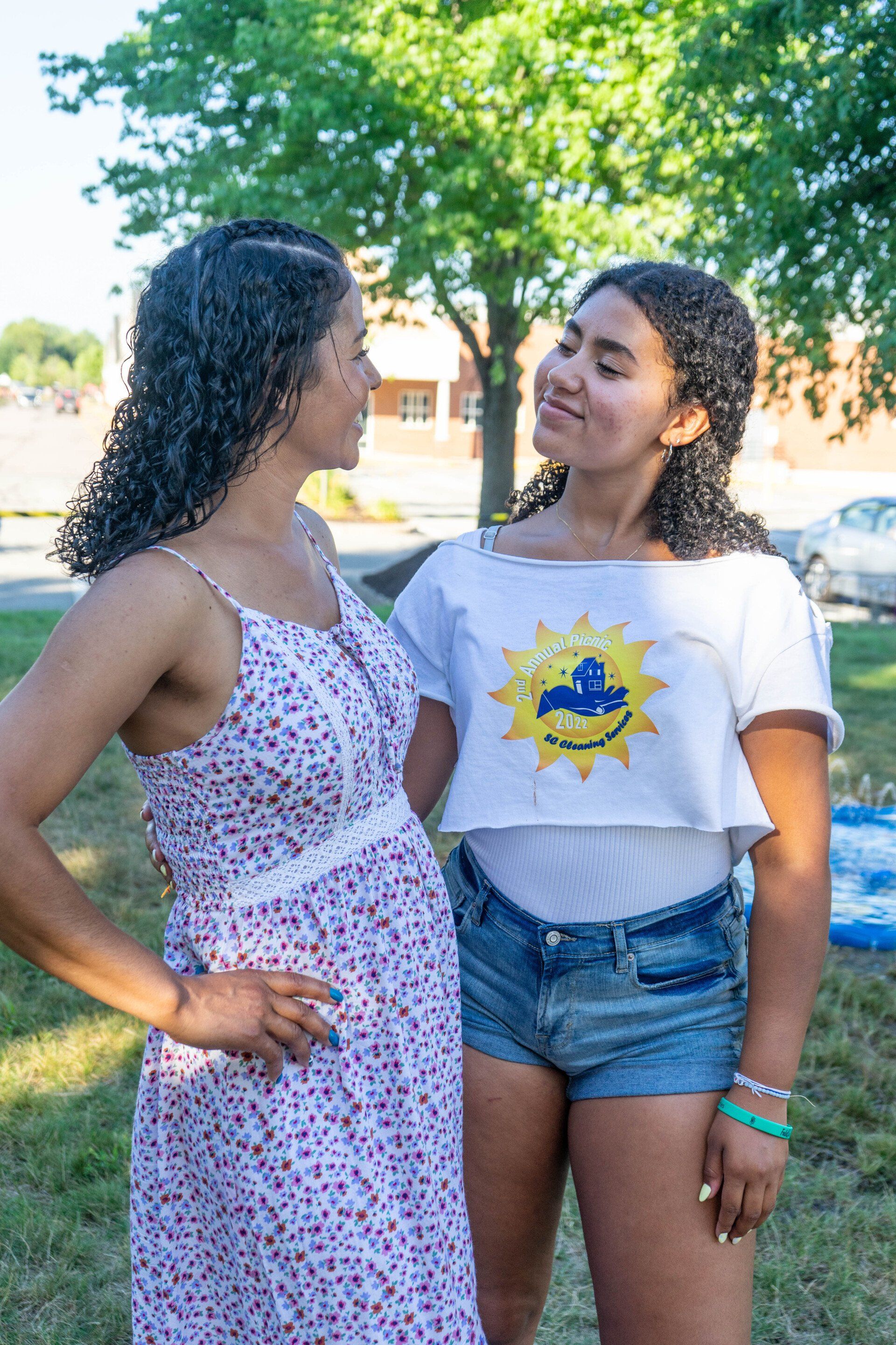 Two women standing outside. One wears a floral dress; the other, a crop top and shorts. They are looking at each other.