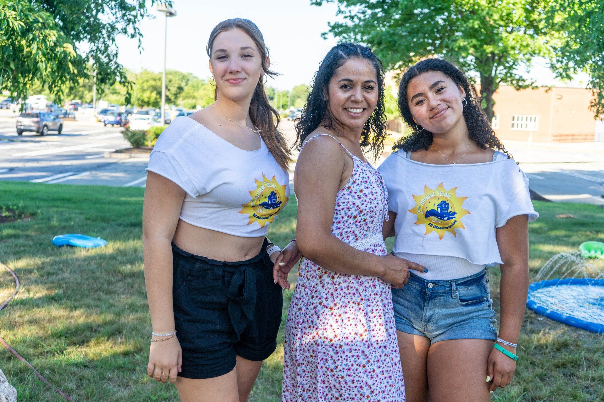 Three women smiling in a park; two wear white crop tops, one in a floral dress.