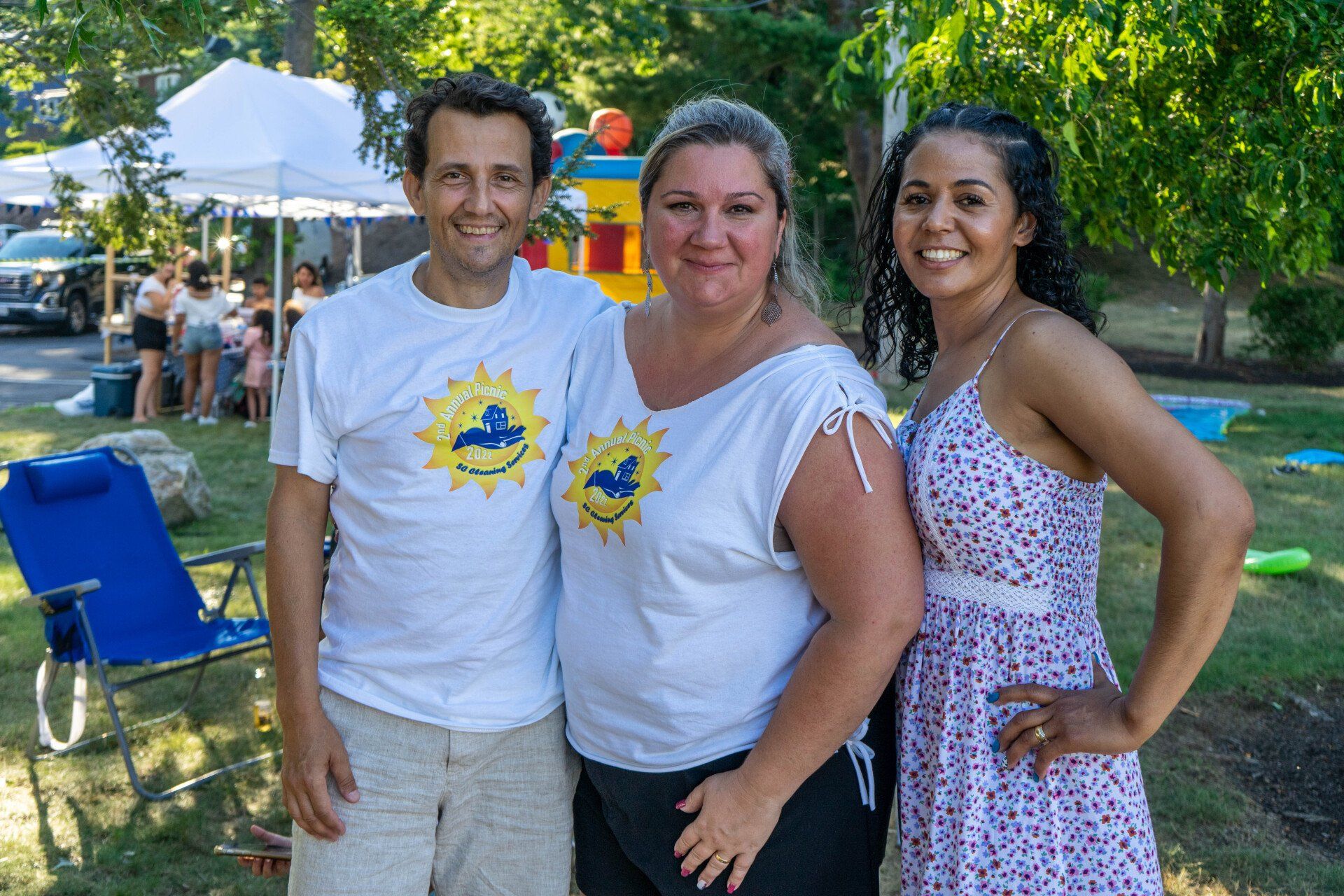 Three people smiling outdoors at an event, two in white shirts with a logo, one in a floral dress.