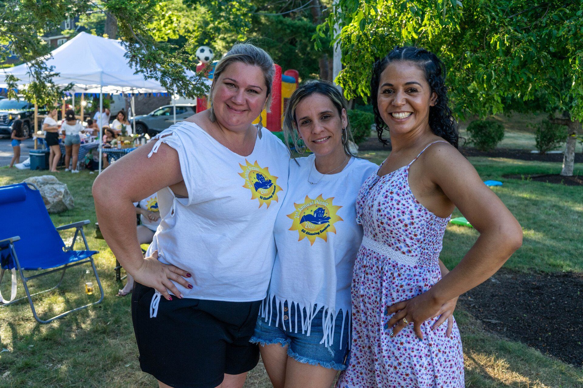 Three women smiling outdoors, wearing white shirts with a sun logo. Green grass and trees in the background.