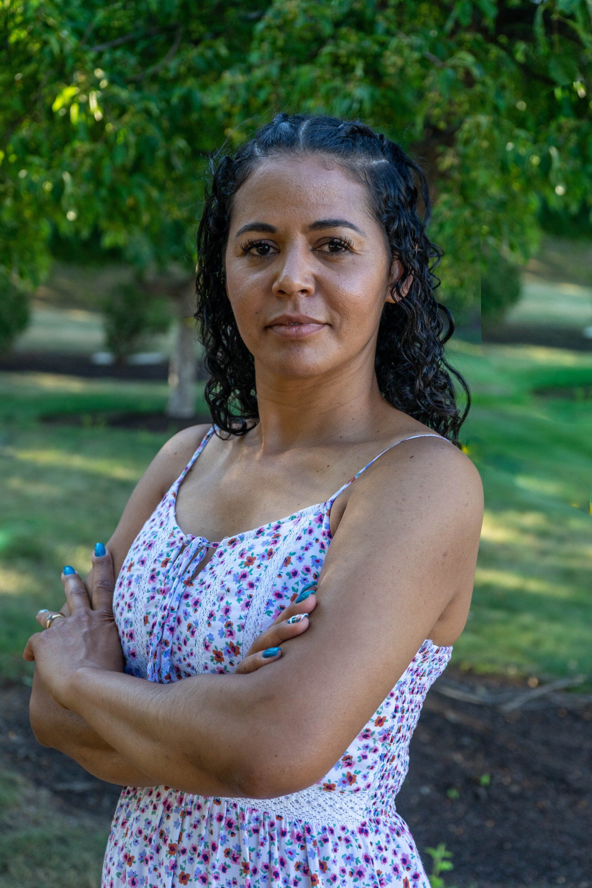Woman with arms crossed wearing a floral dress, outdoors with green trees in the background.