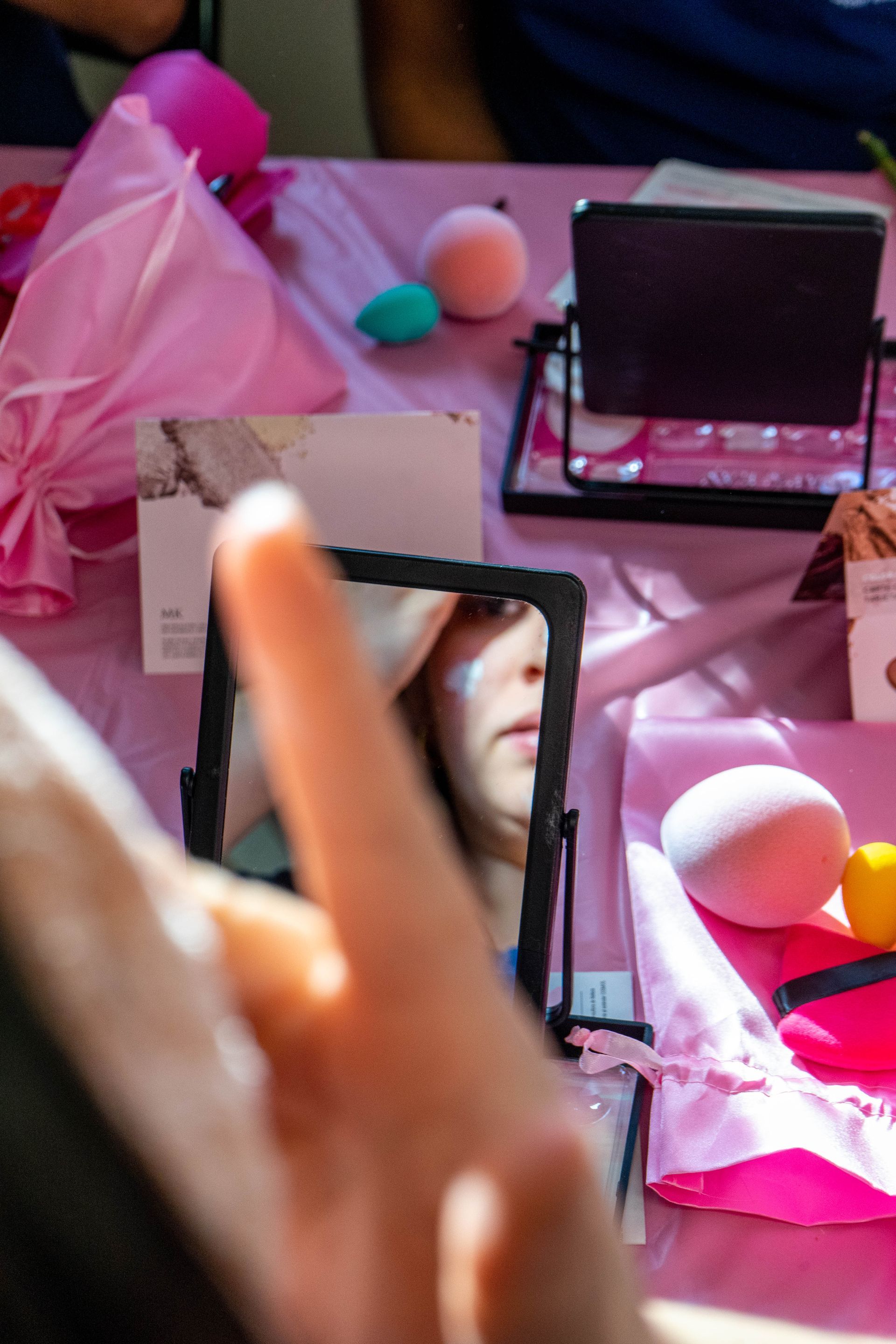 Person applying makeup in a small mirror at a pink table with beauty products.