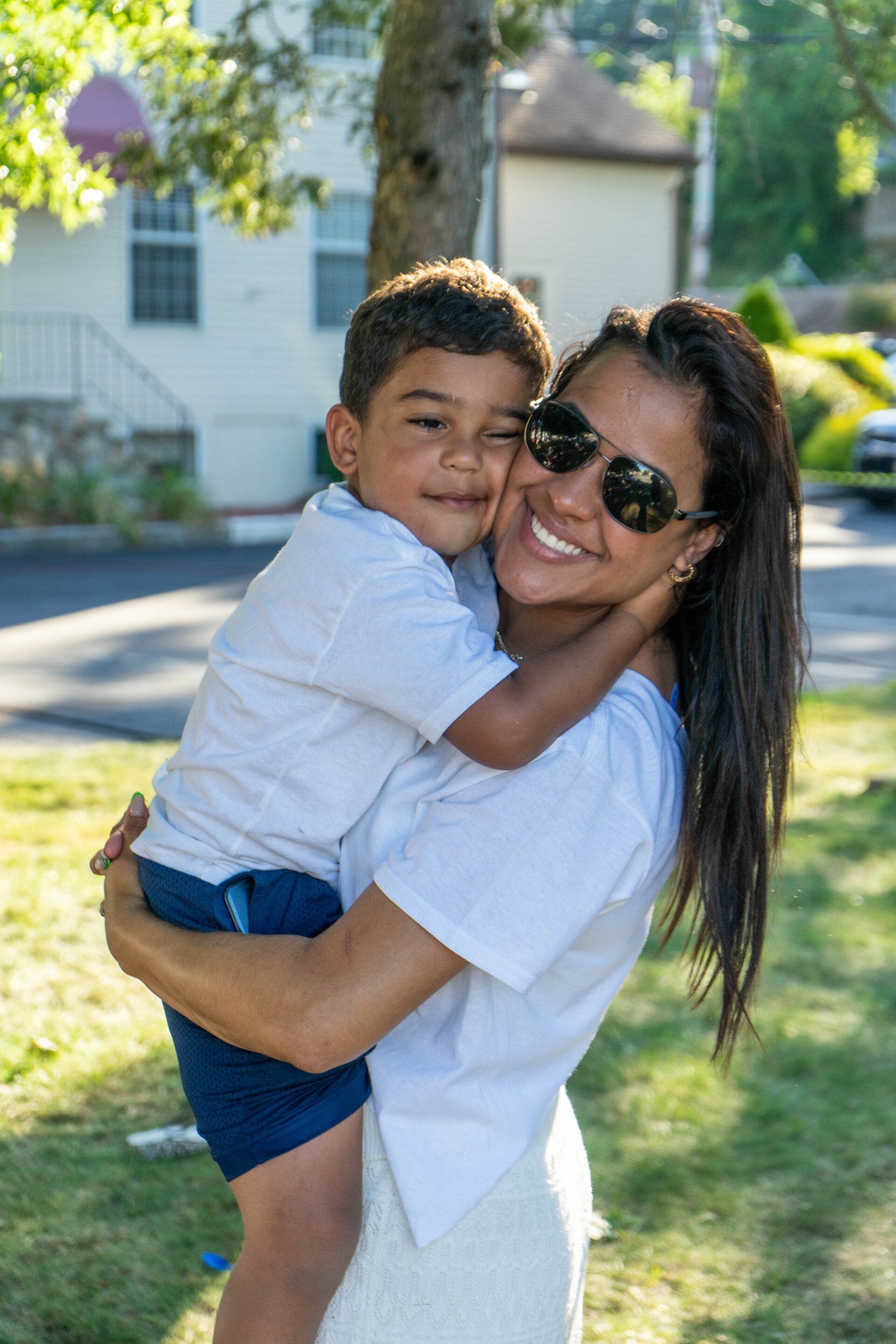 Woman in sunglasses hugs a smiling young boy outdoors, both in white shirts.