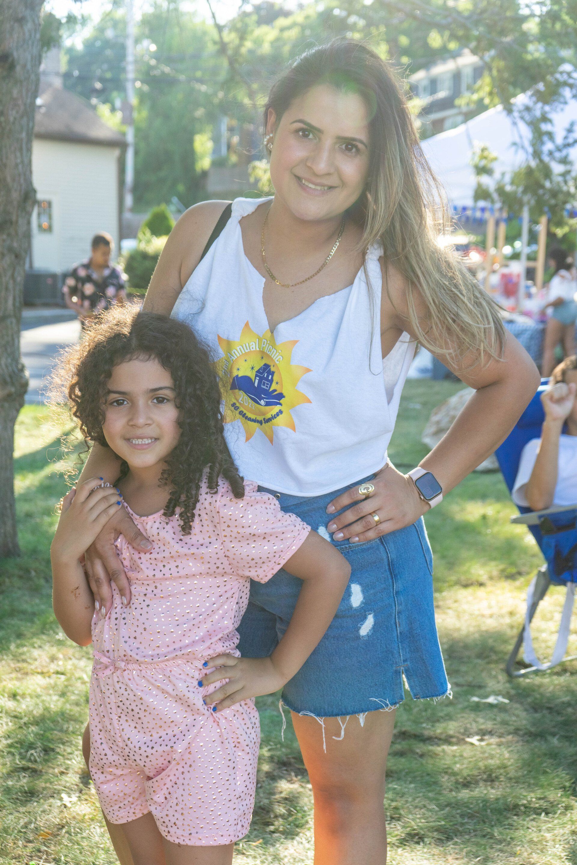 Woman and child smiling, posing outdoors. The woman wears a white tank top and denim skirt. The child wears a pink jumpsuit.