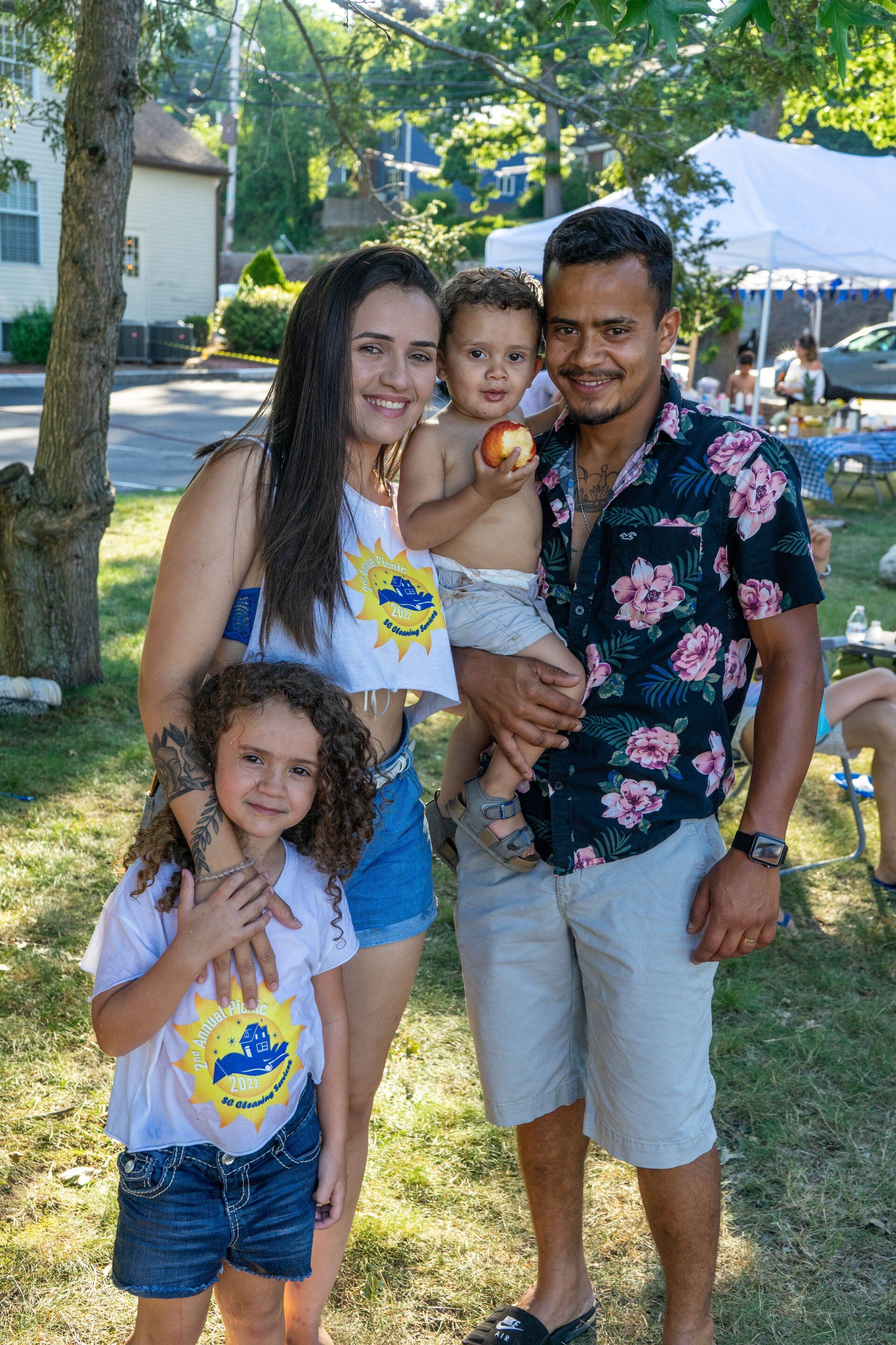 Family of four smiling outside; woman with two children, man in floral shirt.