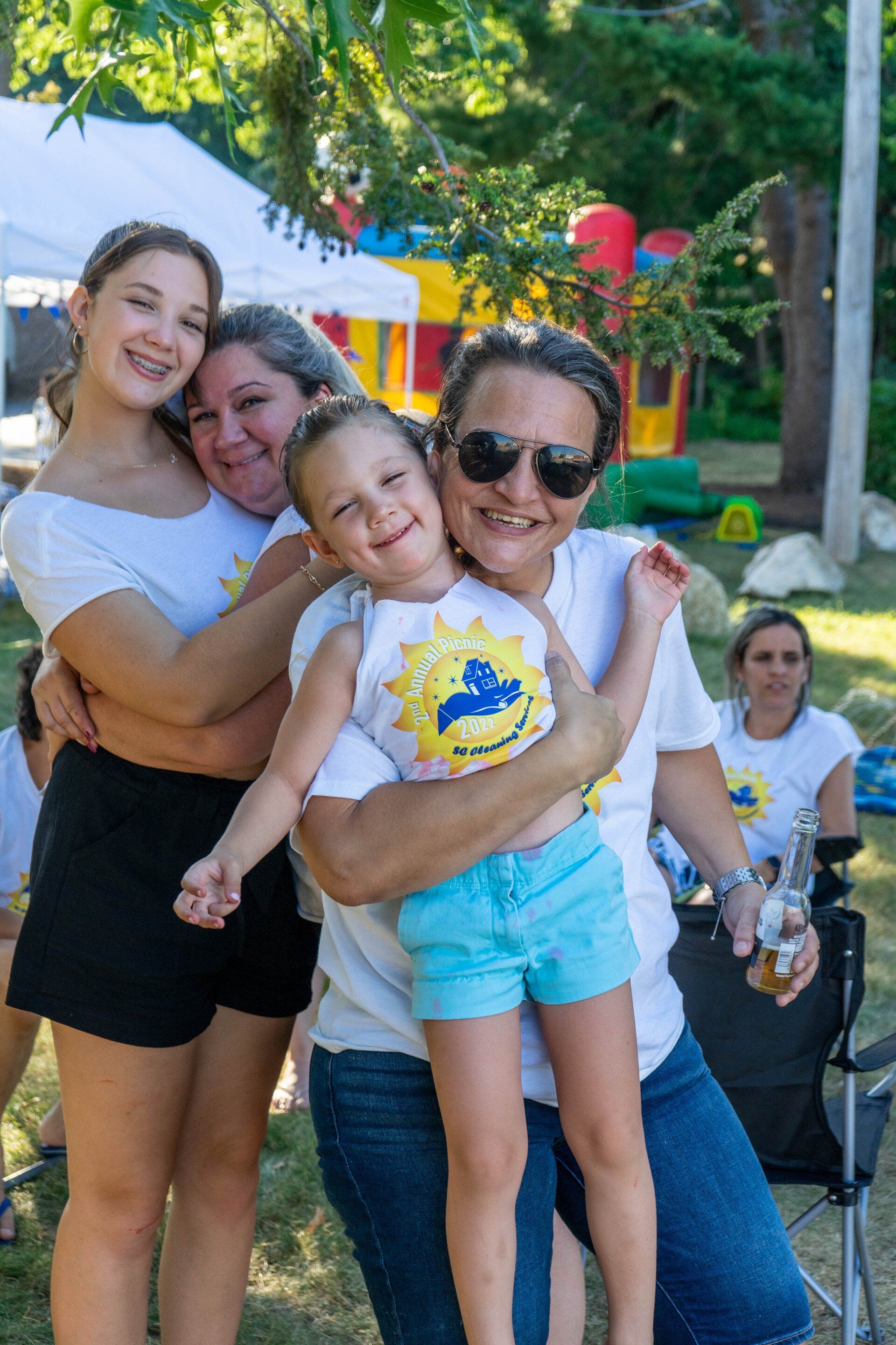 Four smiling people hugging outdoors, wearing matching white shirts with a logo.