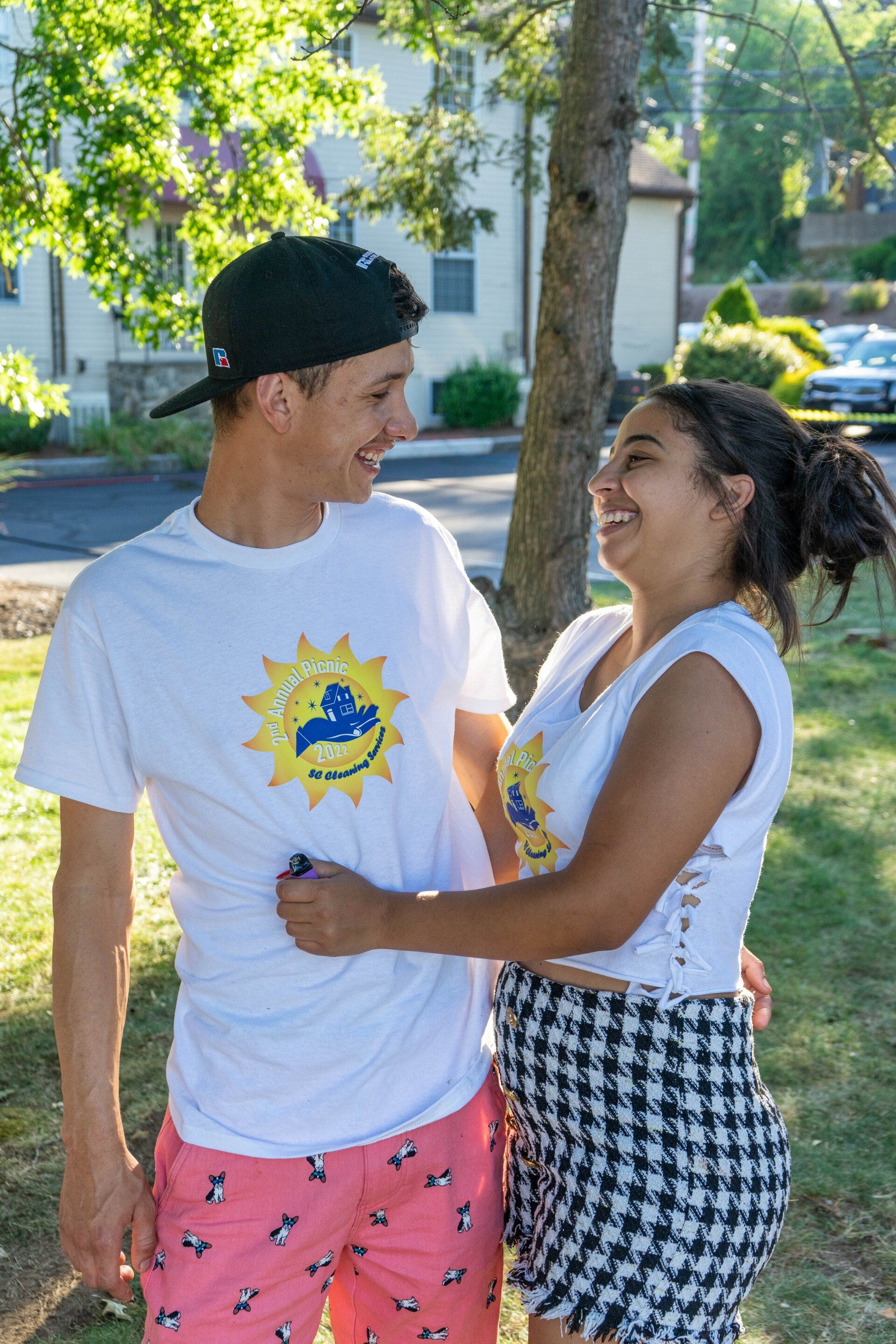 Man and woman laughing, hugging outdoors, both wearing white t-shirts with logo, she in black/white skirt, he in pink shorts.
