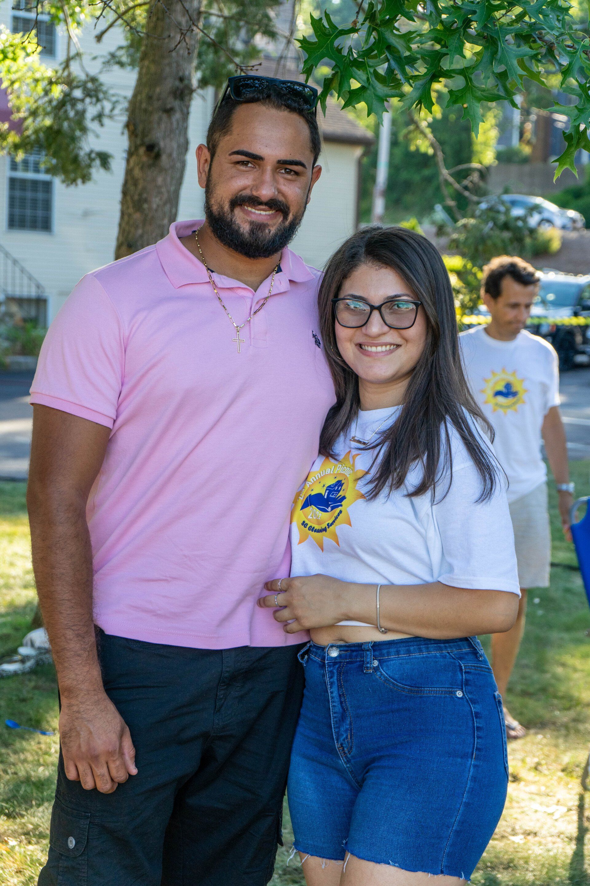 Couple posing for photo outdoors; man in pink shirt, woman in denim shorts, both smiling.