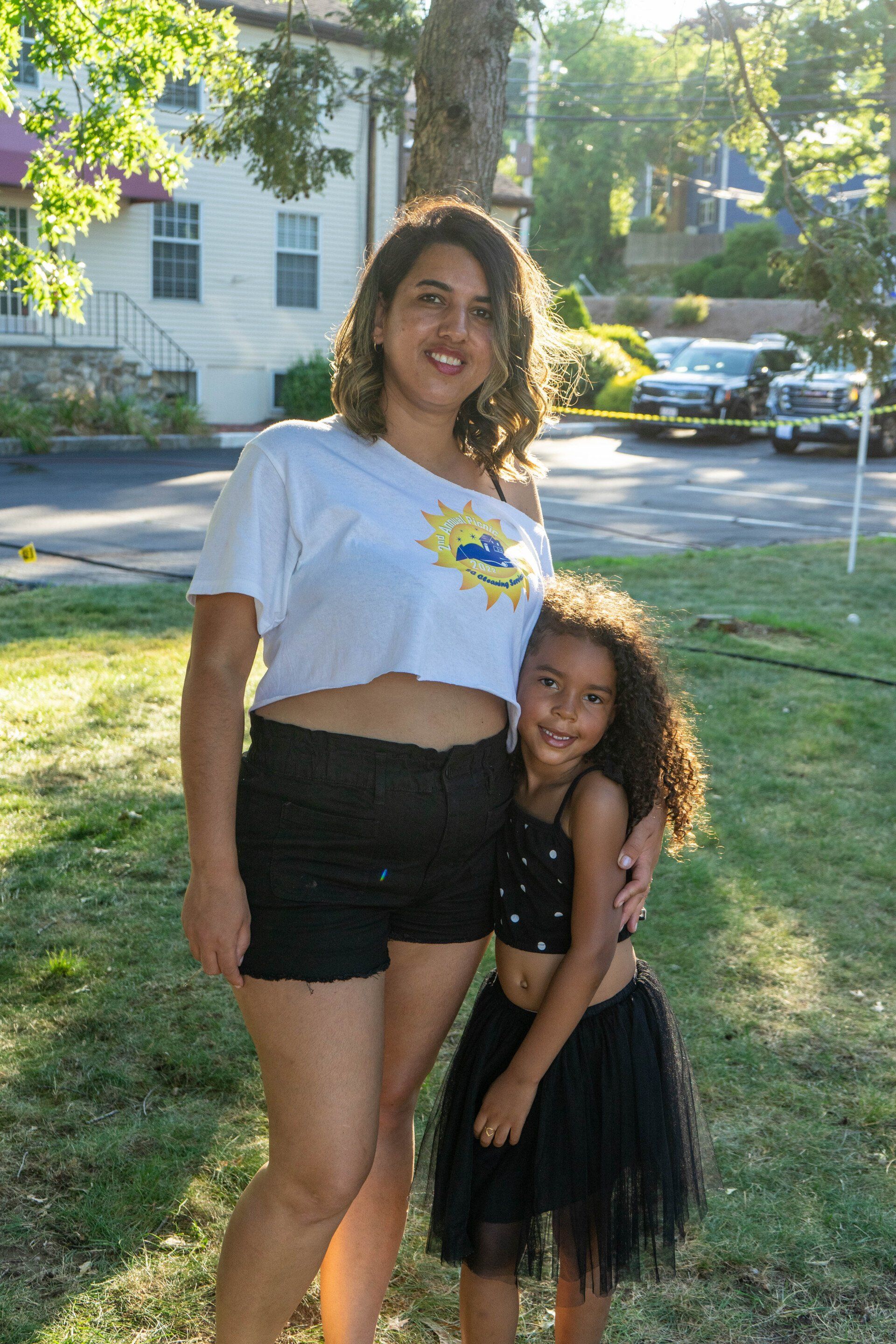 Woman and young girl smiling, hugging outdoors in sunlight. Woman in white shirt and black shorts; girl in black top, skirt.
