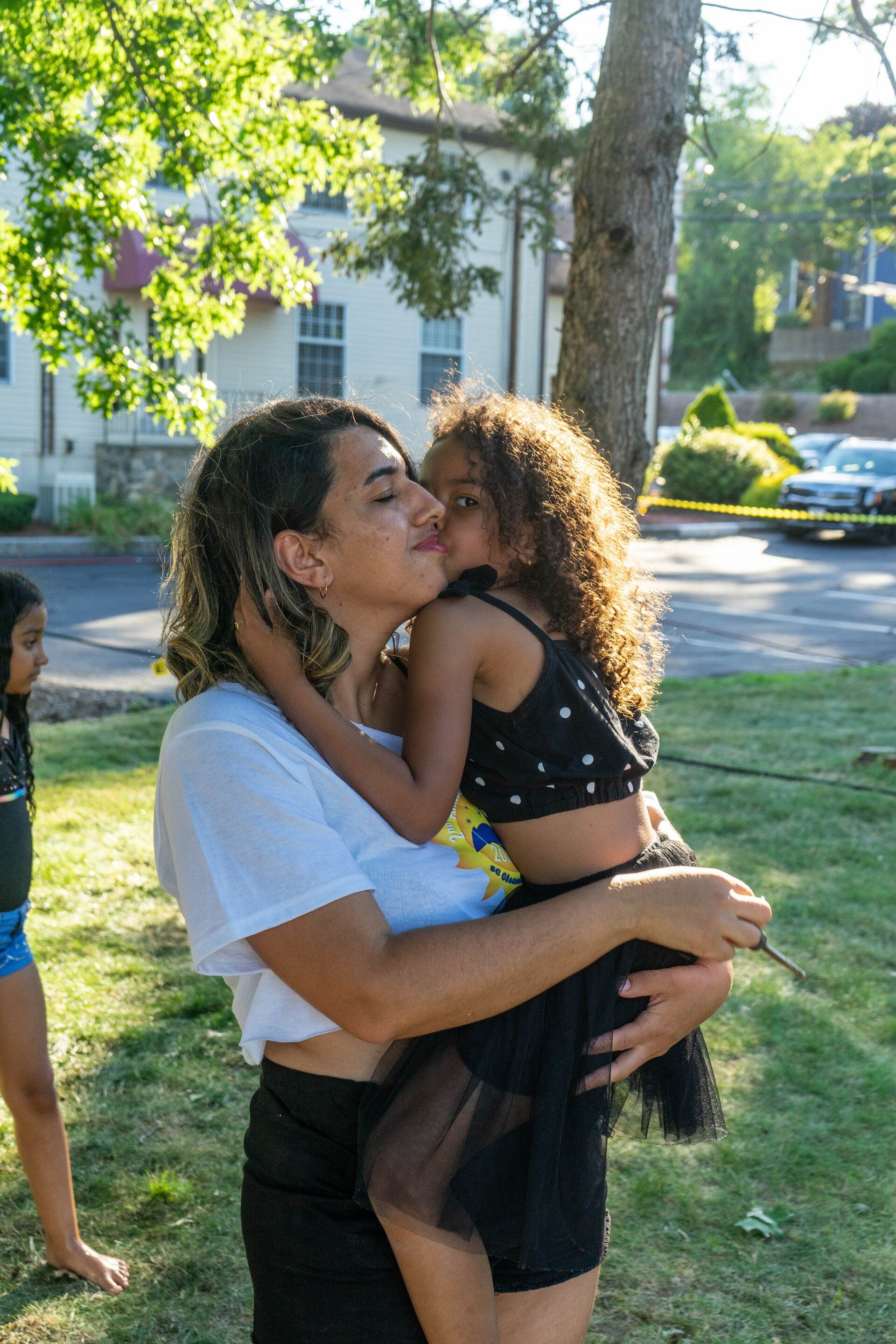 Woman holds and kisses a young girl outdoors, green grass, tree in background, sunny day.